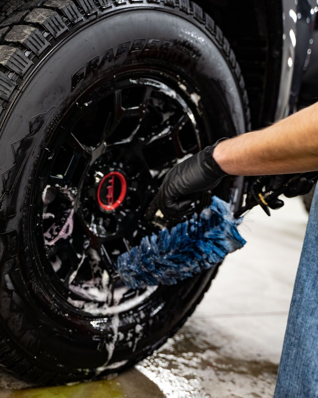 A person in black gloves cleans a black car wheel with a blue brush, using soapy water in a garage setting.