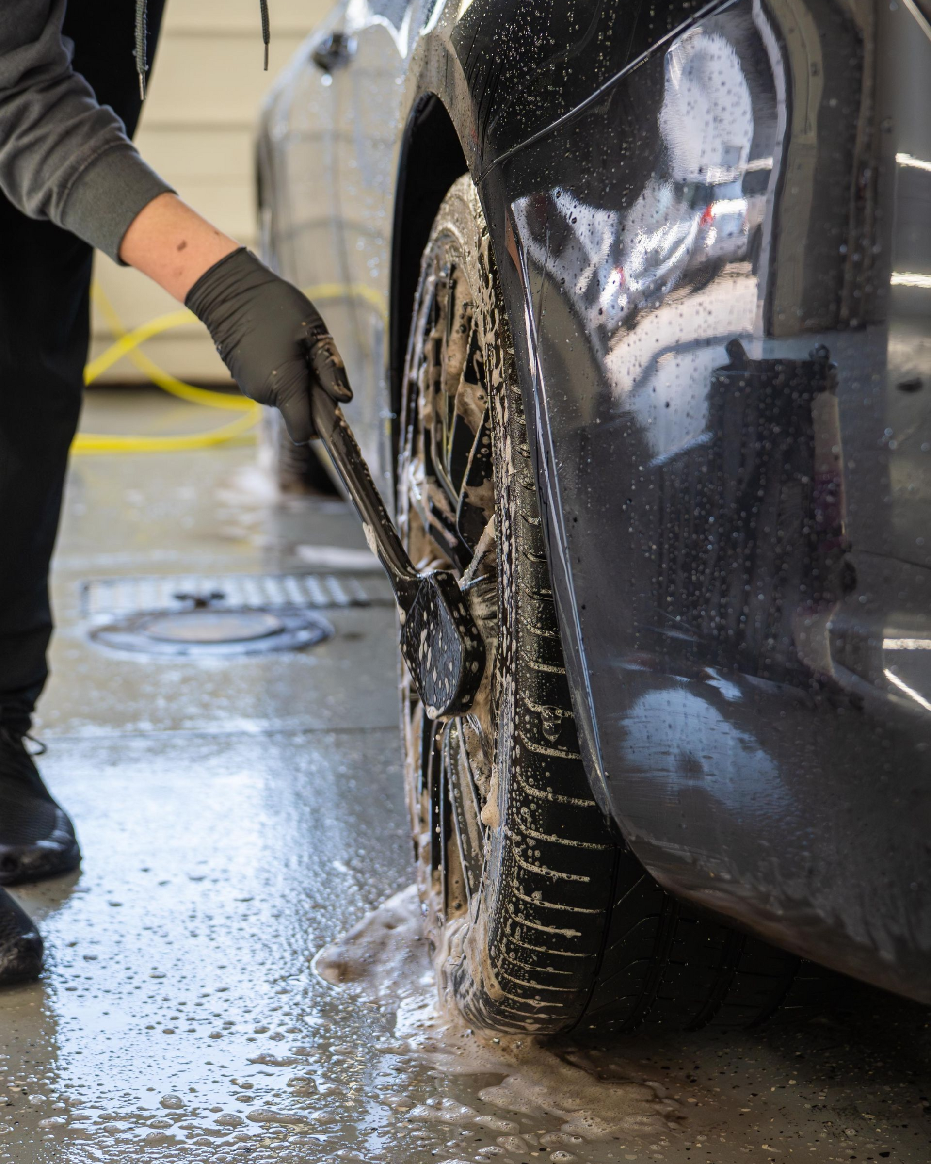 A person wearing a black glove uses a brush to scrub dirt and soap off a car wheel in an outdoor wash bay.