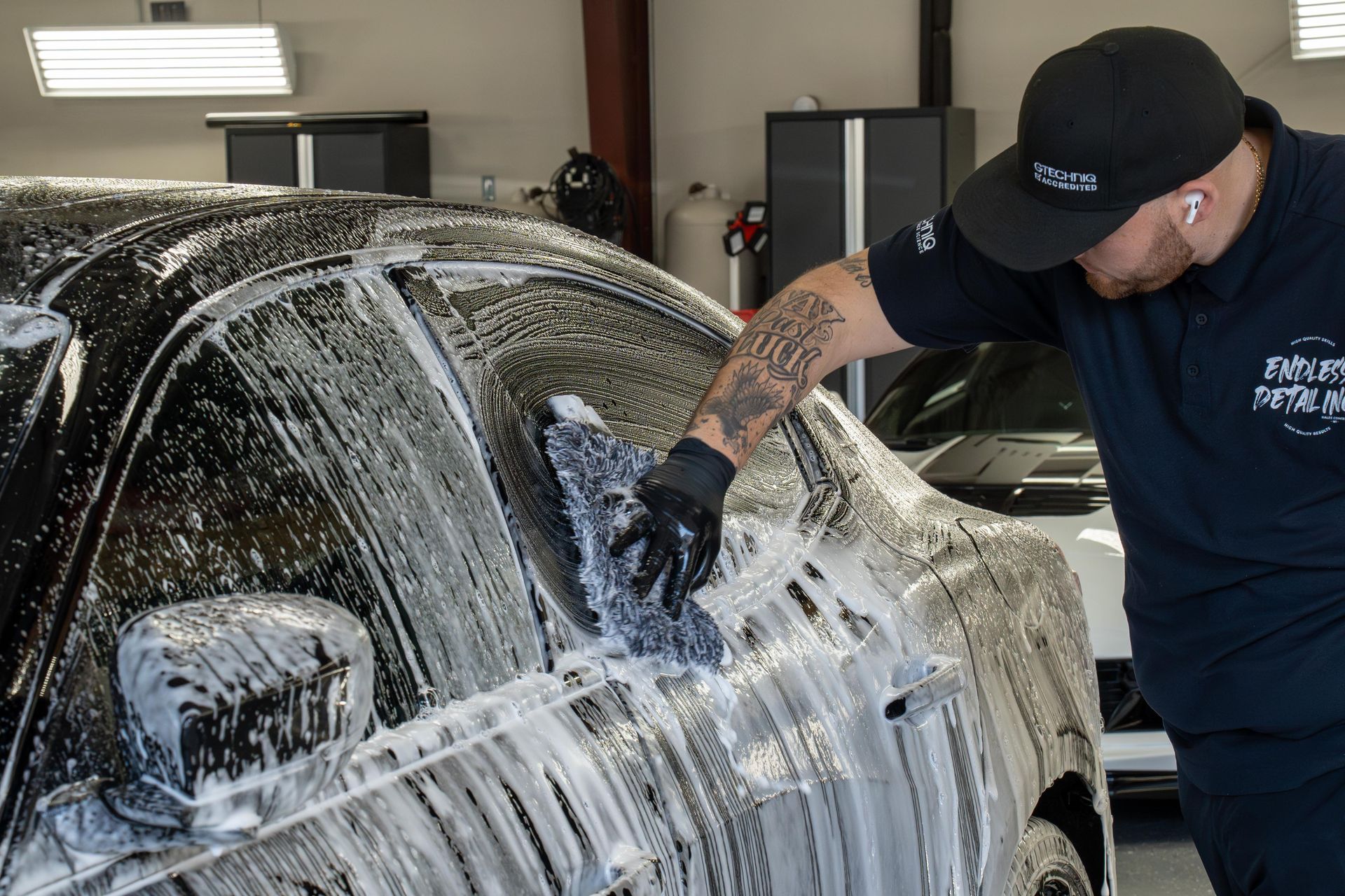 A detailer in a black uniform and cap uses a foam-covered mitt to wash a soap-covered car in a garage.