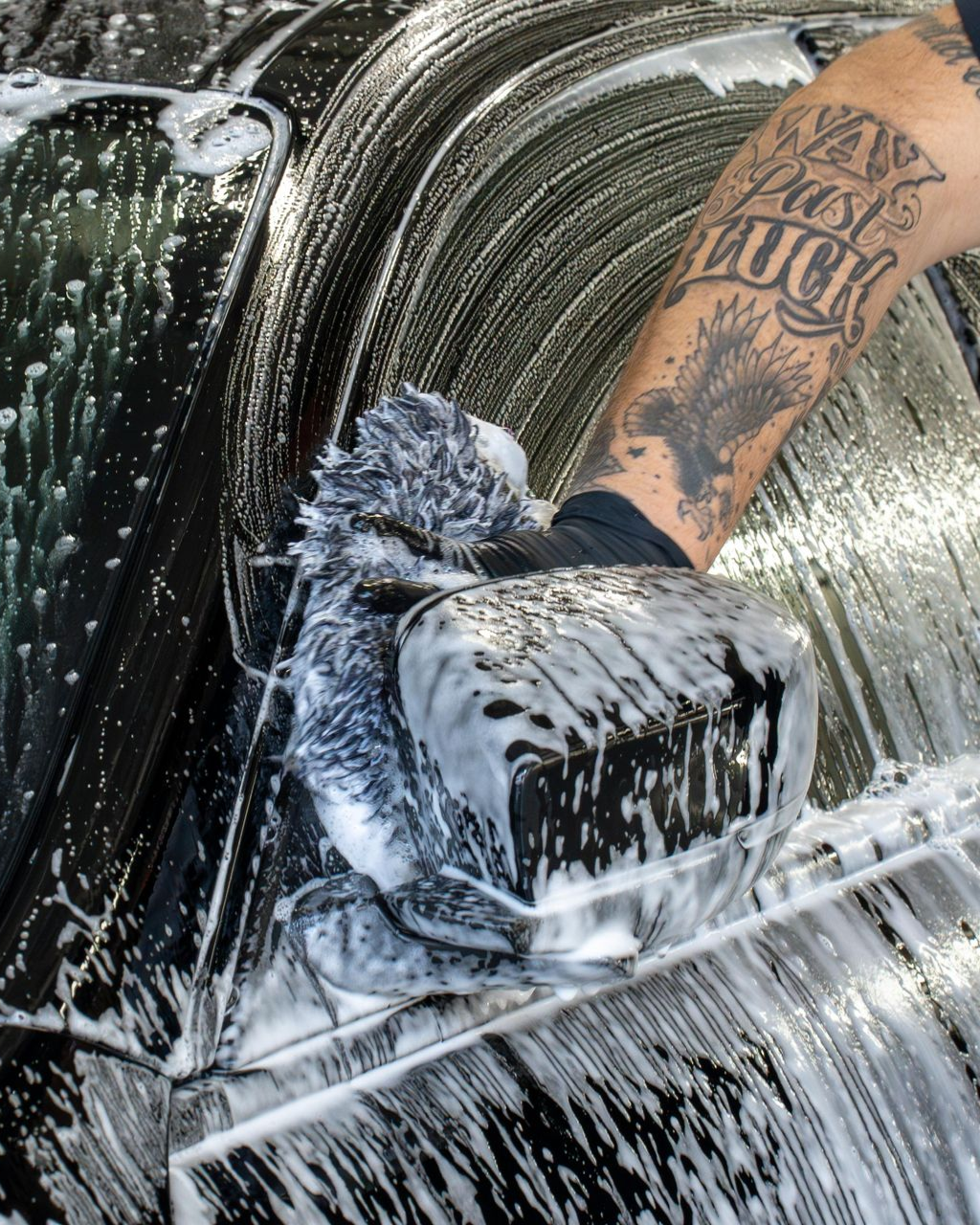A gloved hand uses a grey mitt to wash the soap-covered side mirror of a black car.