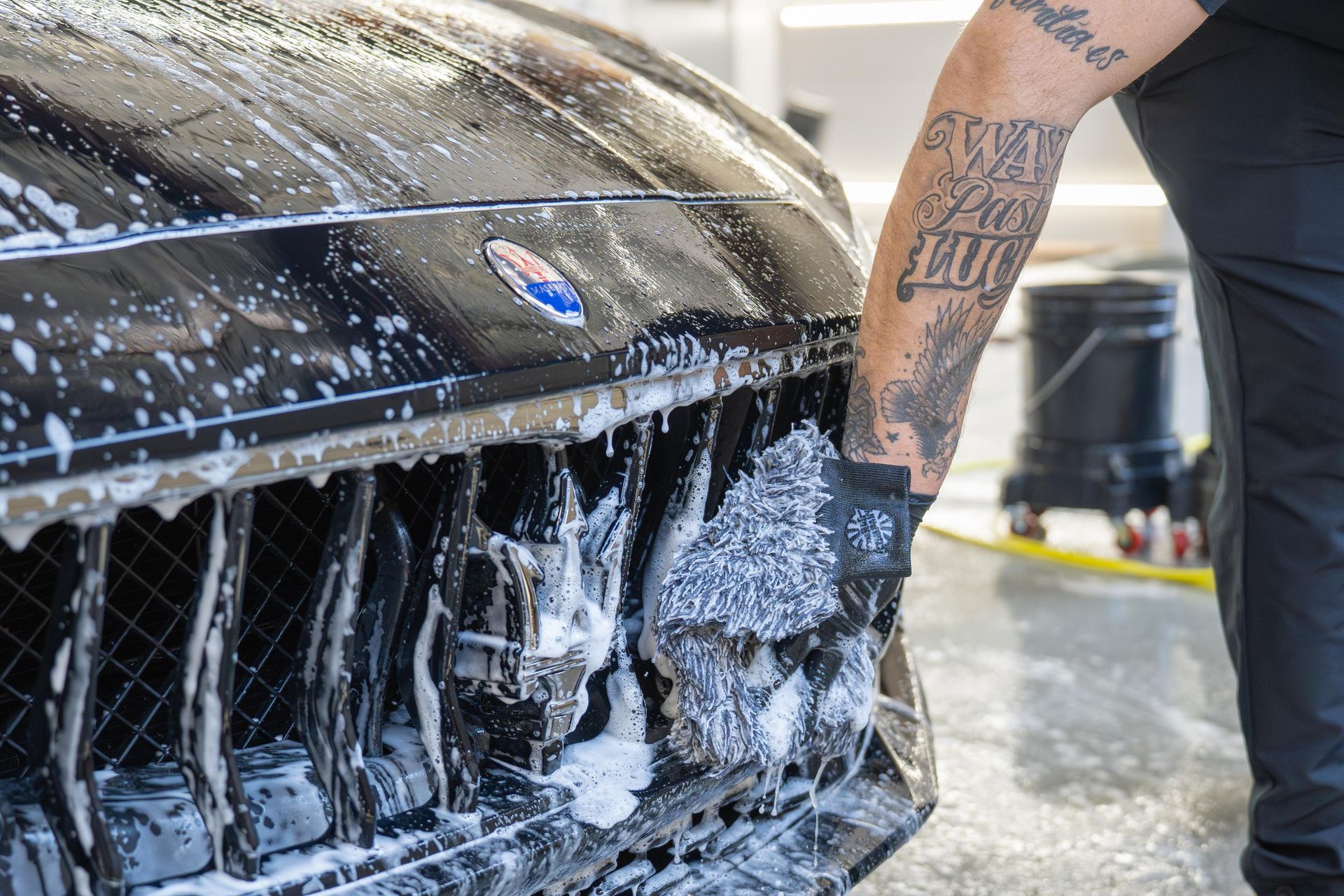 A person with a tattooed arm uses a sponge to wash the front grille of a black Maserati covered in soap suds.