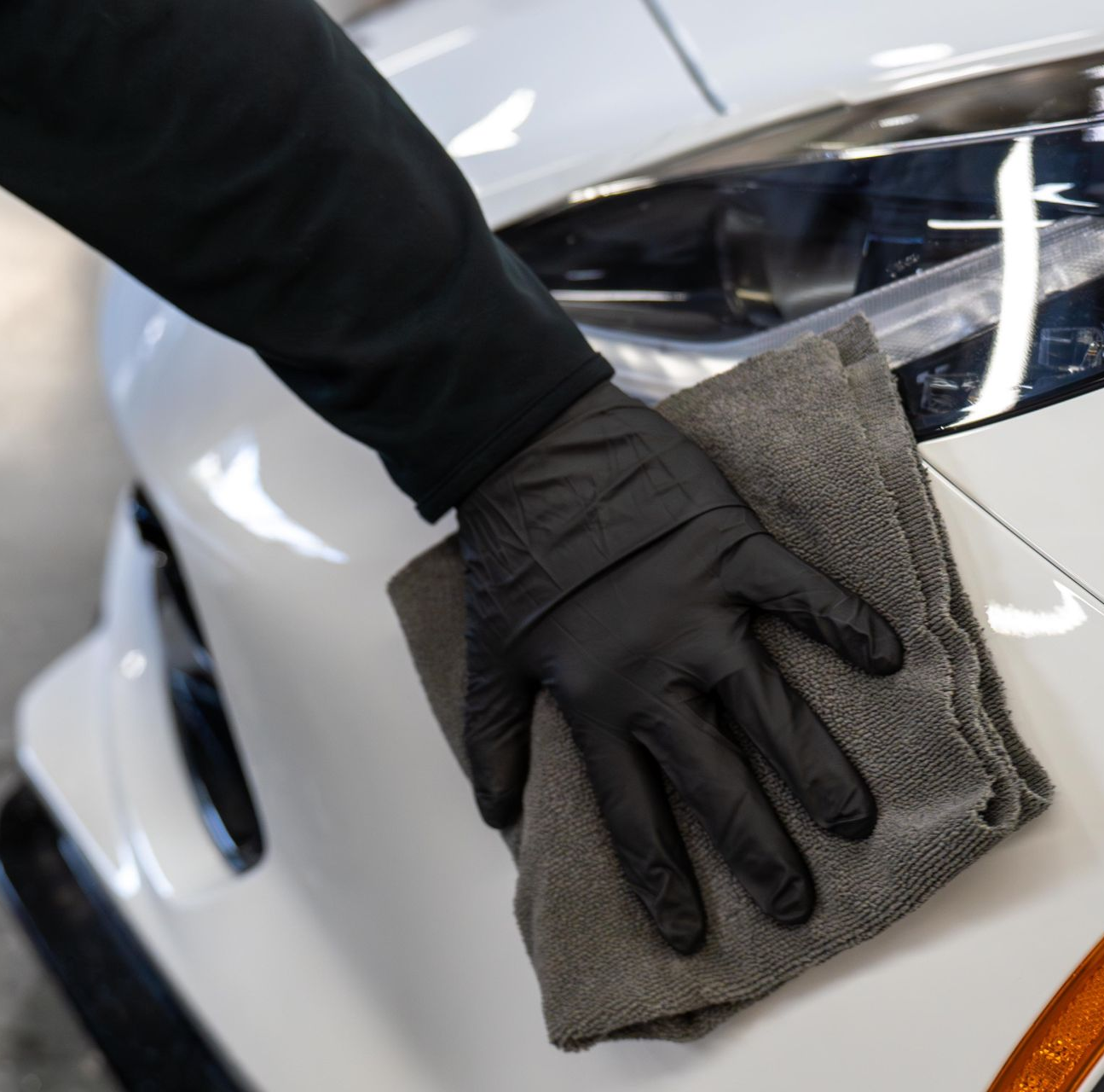 A person wearing black gloves wipes a white car with a gray microfiber cloth.