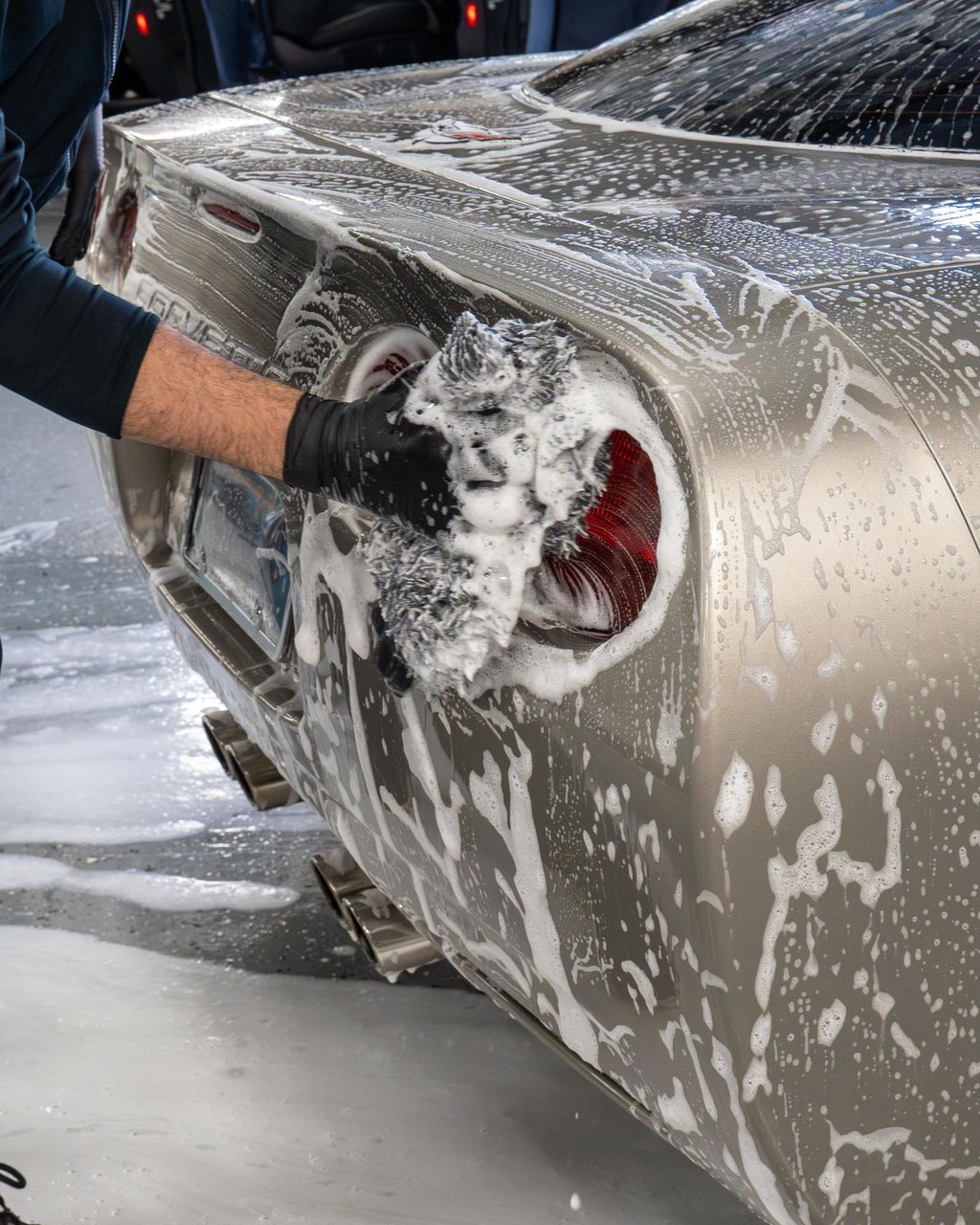 A person wearing black gloves washes the rear taillight of a champagne-colored car with soapy foam.