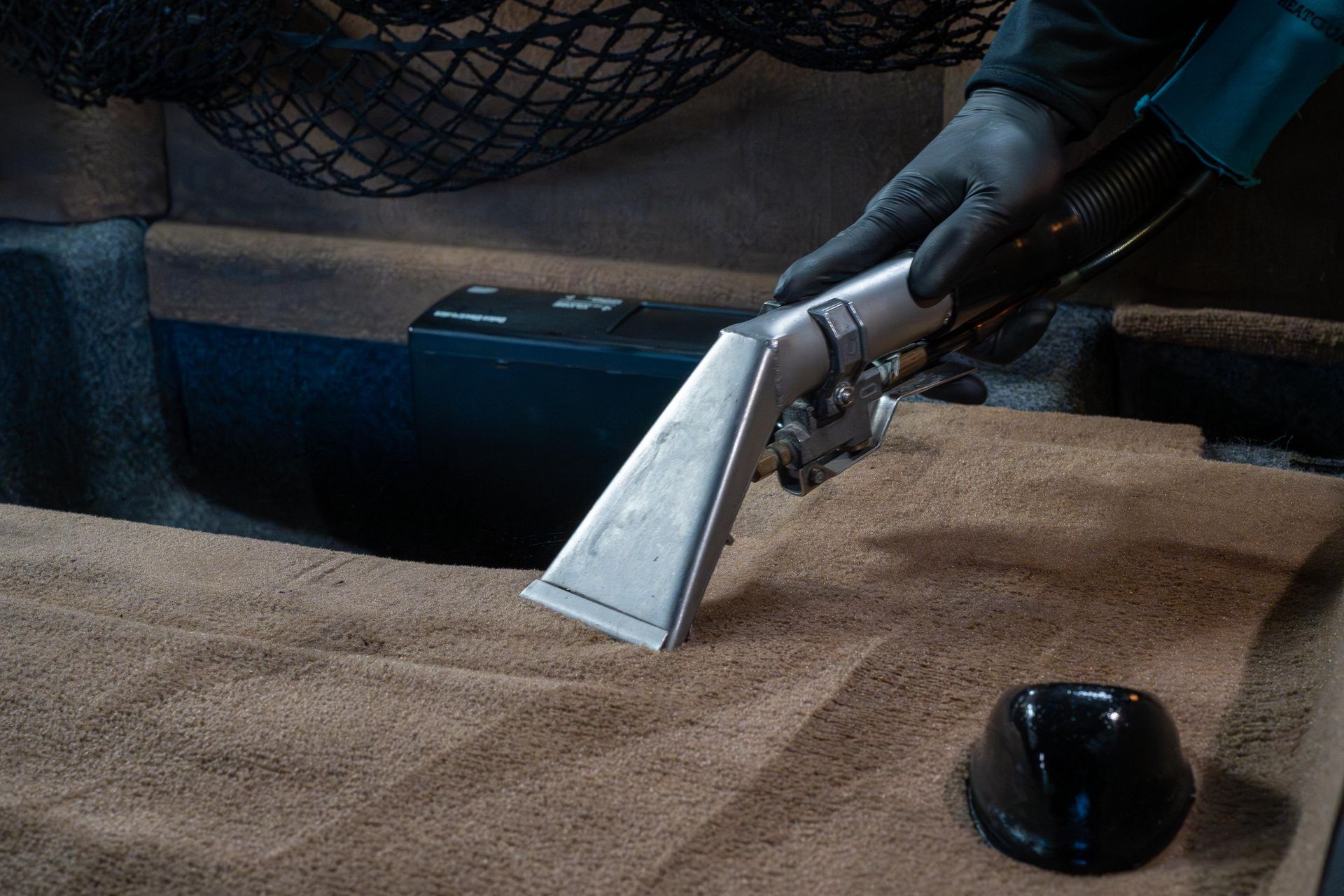 A gloved hand uses a vacuum extraction tool to deep clean tan carpet inside a vehicle.