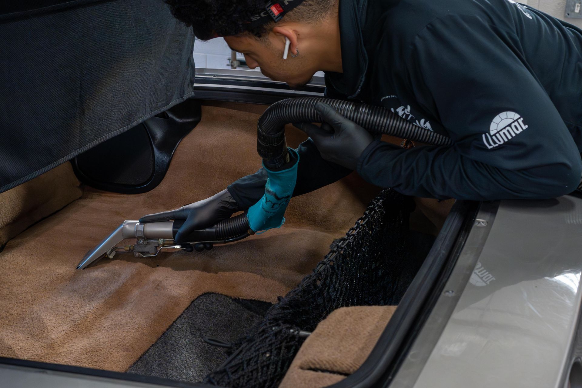 A person in a black shirt and gloves uses a vacuum extraction tool to clean the beige carpet of a vehicle trunk.