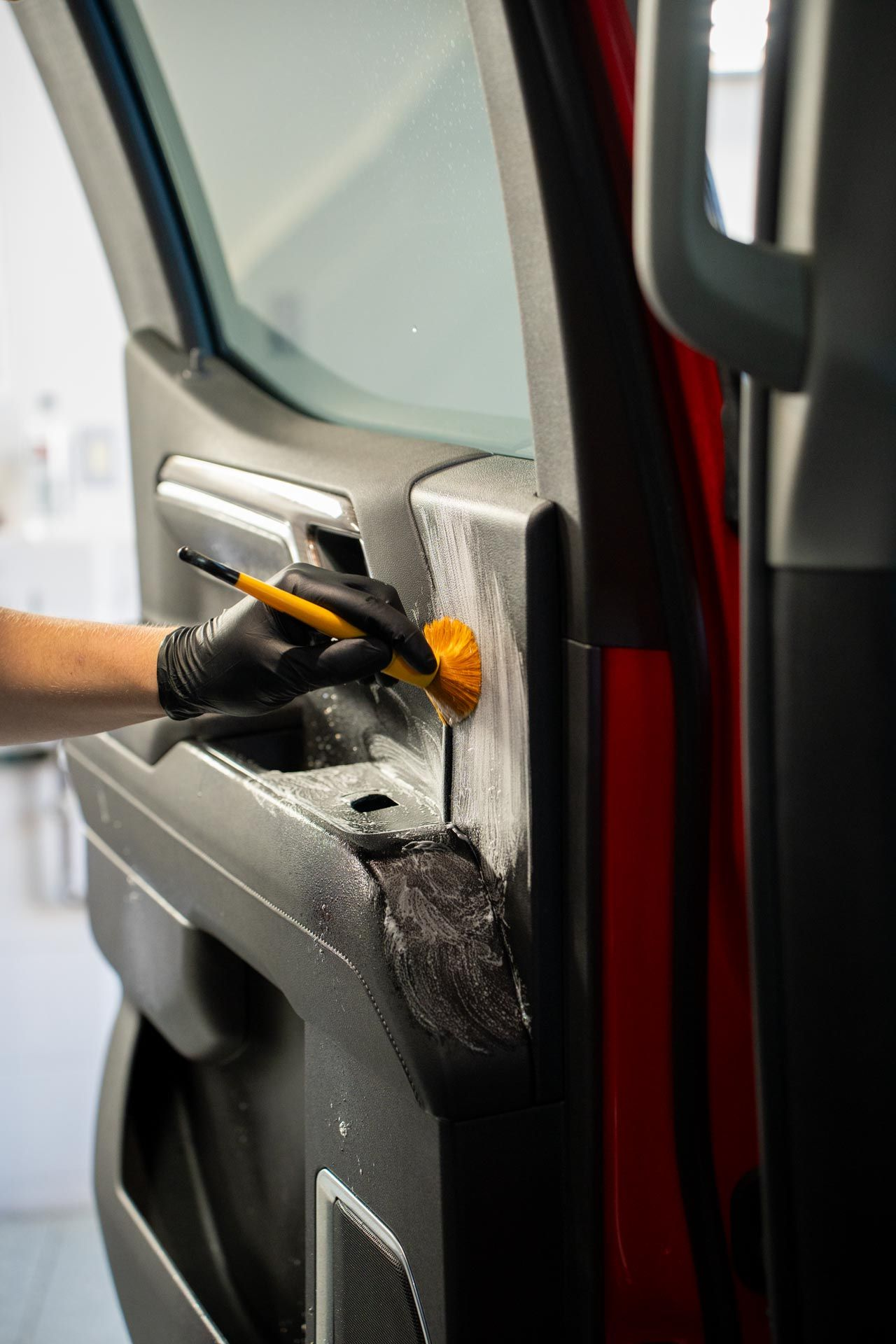 A gloved hand uses a small yellow brush to scrub a soapy cleaning solution into the textured surface of a car door panel.