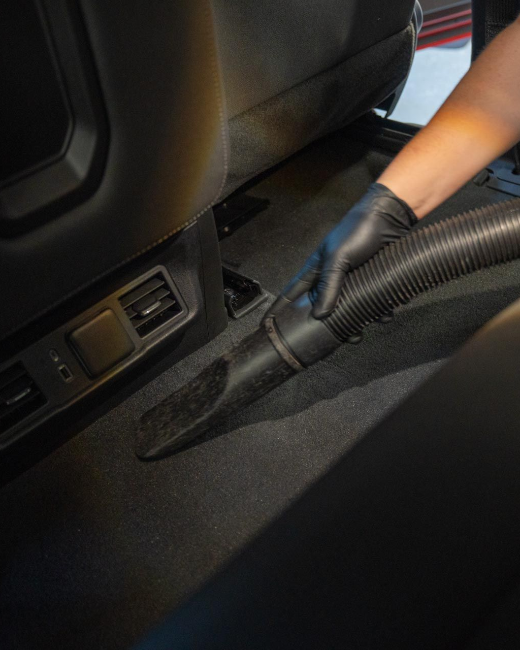 A person wearing a black glove uses a vacuum cleaner nozzle to clean the dark carpet inside a vehicle.