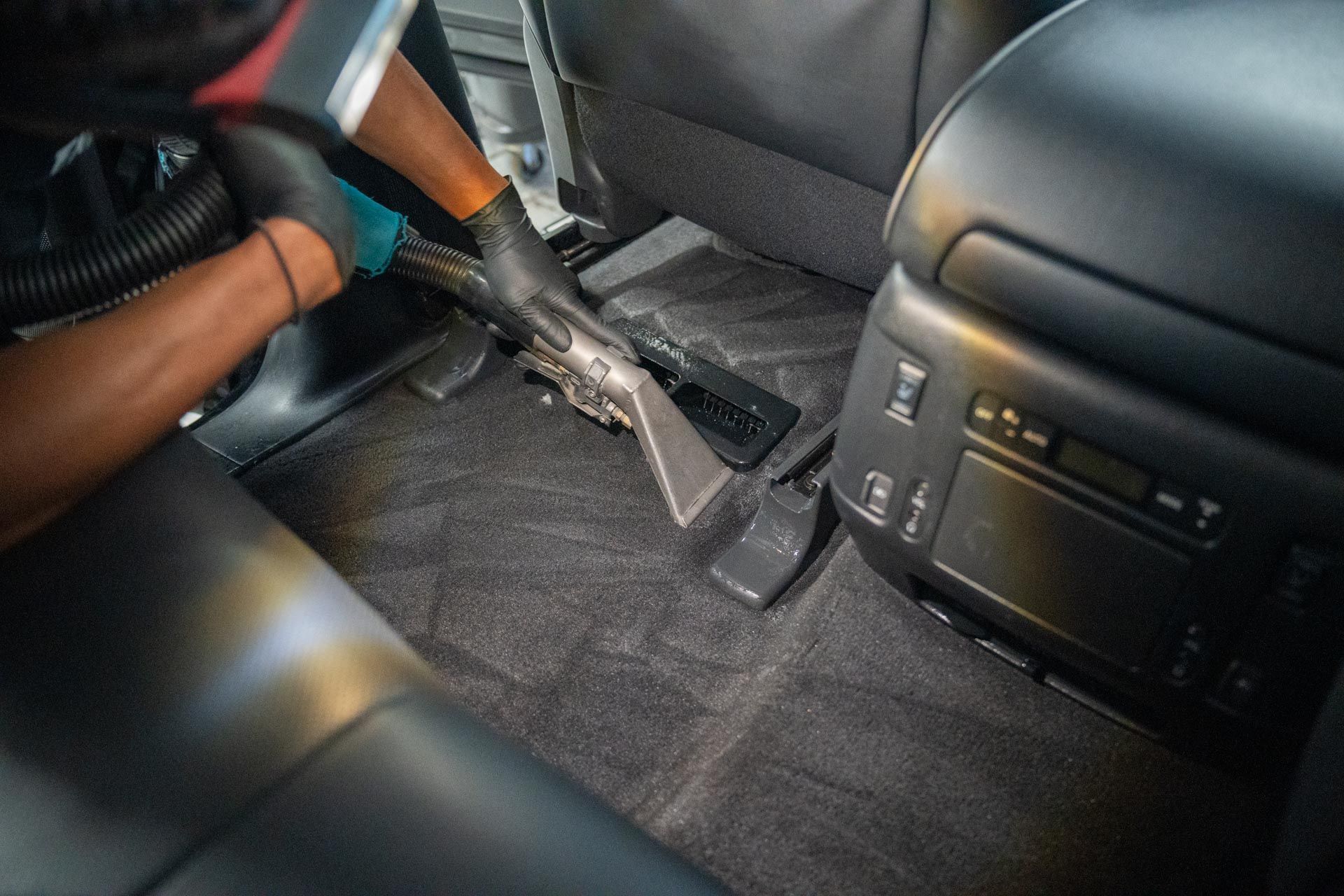 A person wearing black gloves uses an extraction vacuum tool to deep clean the dark carpet of a vehicle interior.