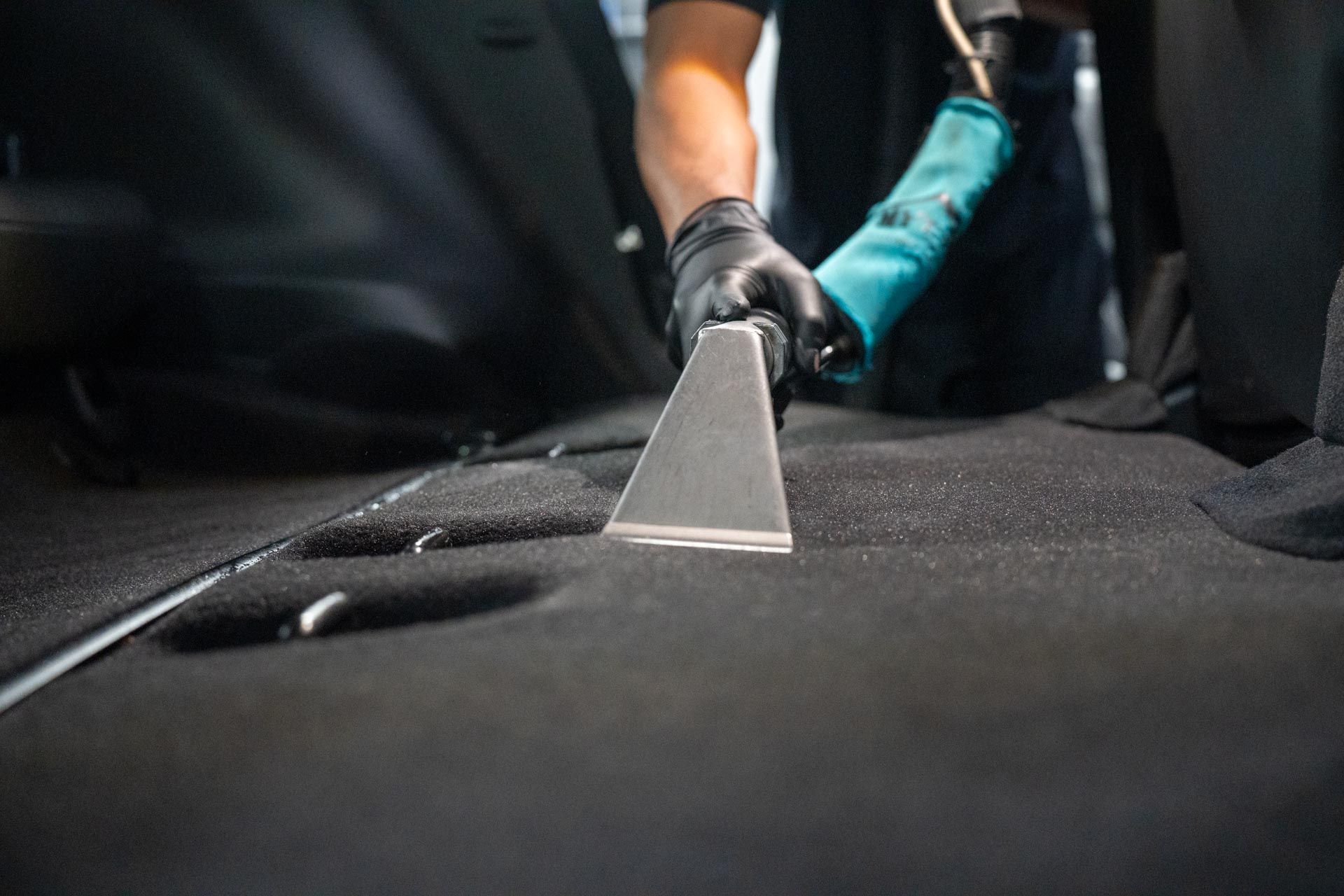 A gloved hand uses a vacuum extraction tool to deep clean a dark fabric car interior.