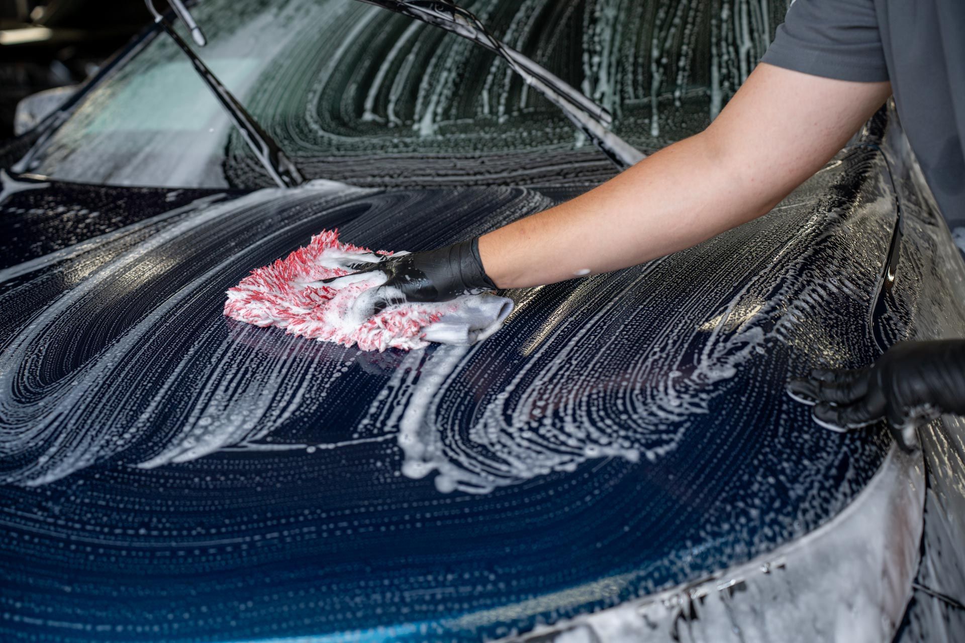A person wearing black gloves uses a white and red wash mitt to clean the soapy, dark blue hood of a car.