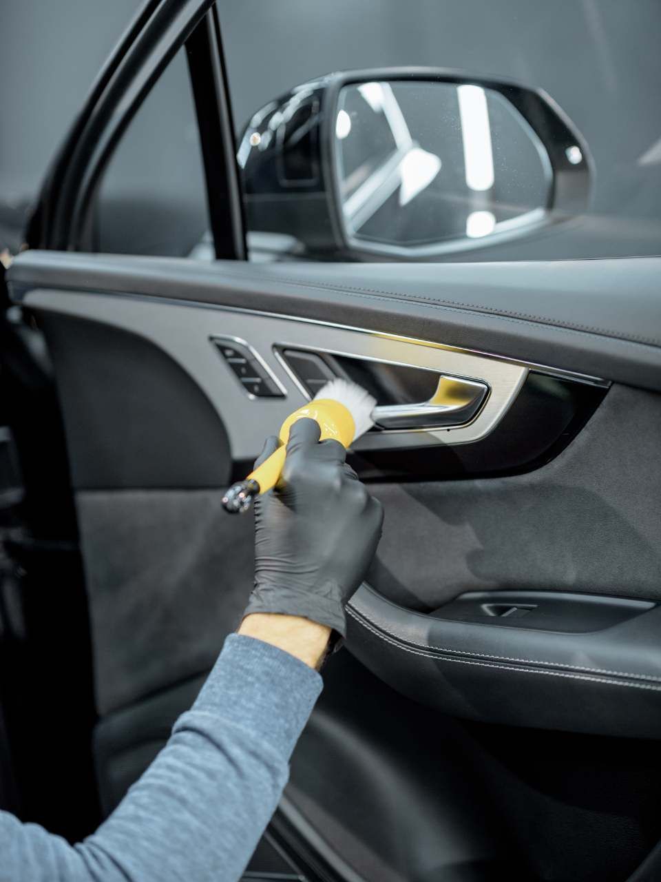 A detailer in a black uniform and cap uses a foam-covered mitt to wash a soap-covered car in a garage.