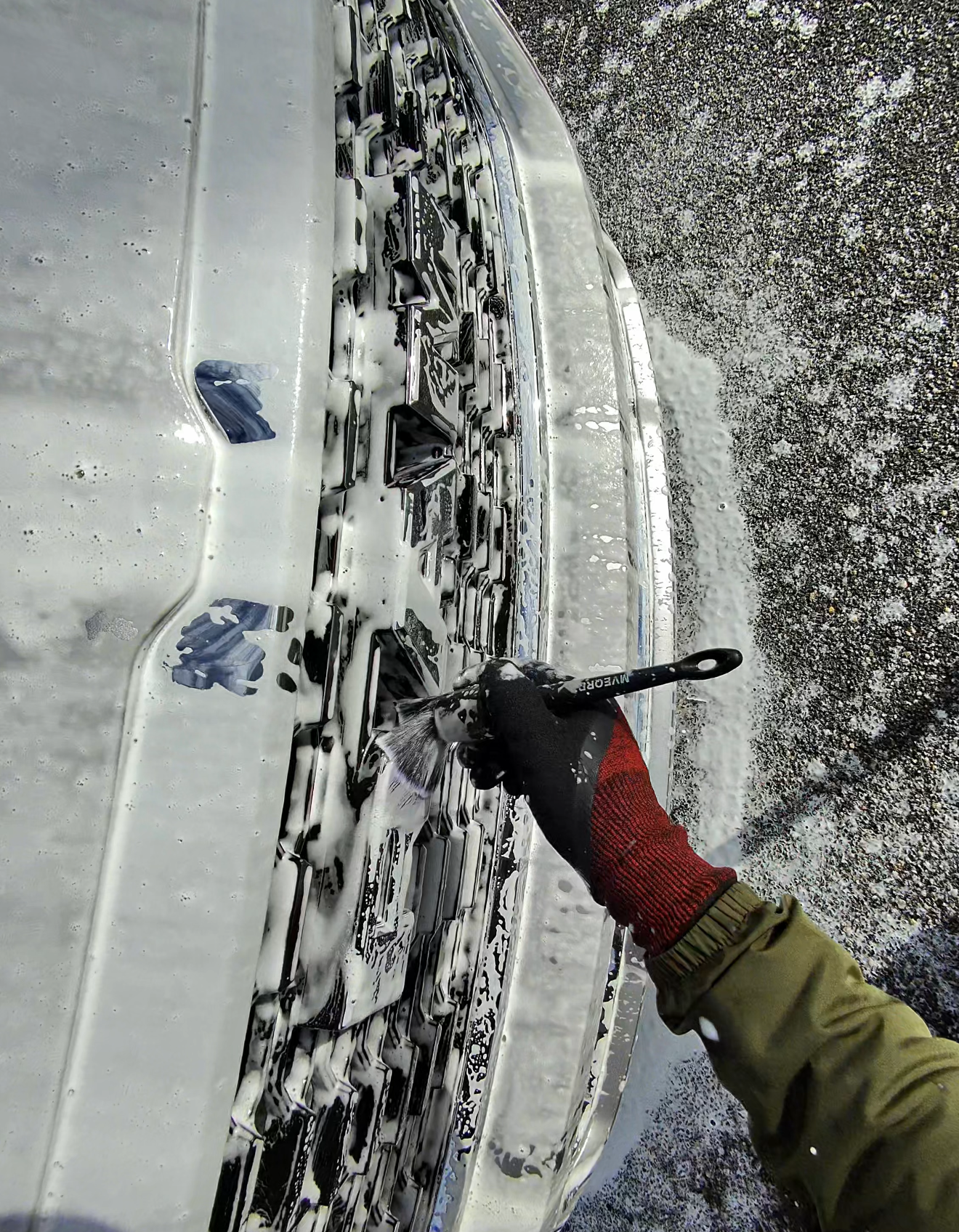 A gloved hand uses a brush to clean soap foam from the front grille of a vehicle.