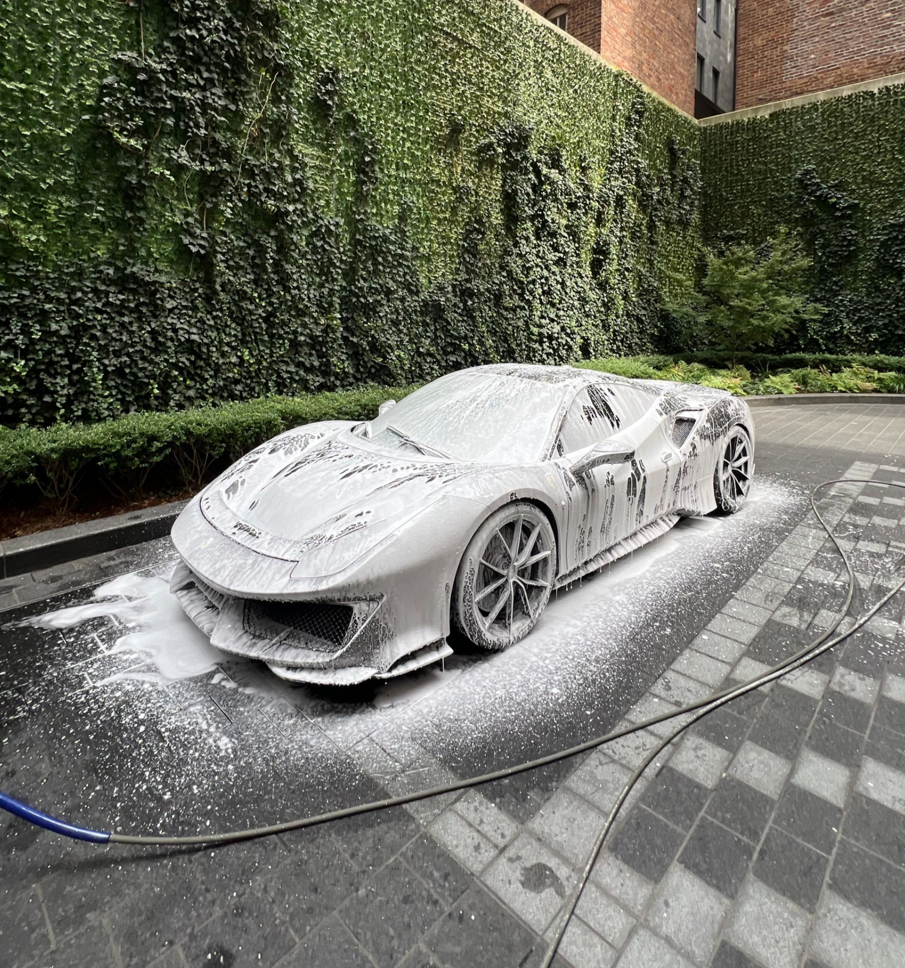 A luxury sports car covered in thick white car-wash foam parked on a tiled driveway next to a tall, ivy-covered wall.