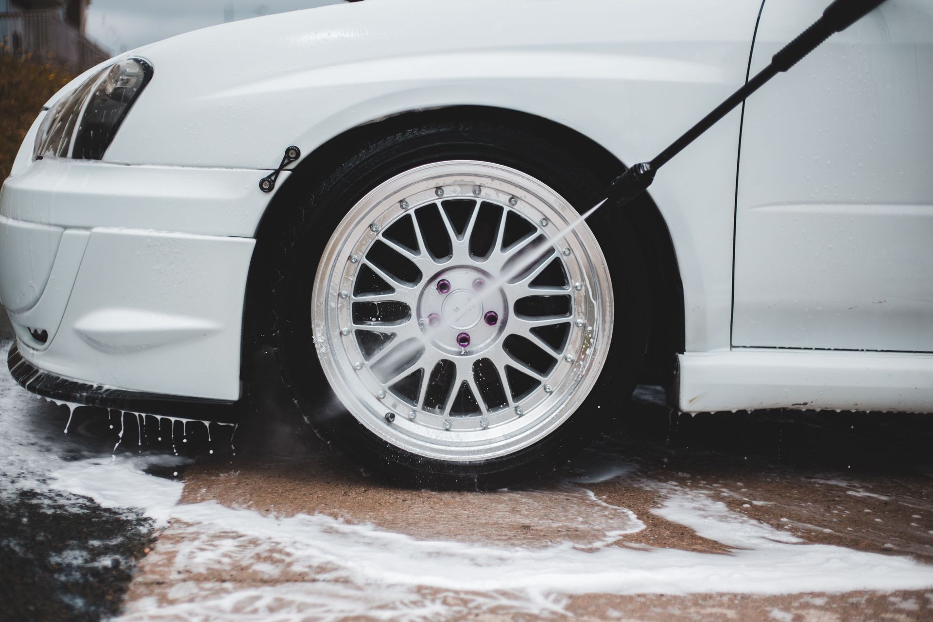 A person uses a pressure washer to clean the tire of a silver car inside a workshop with other vehicles.