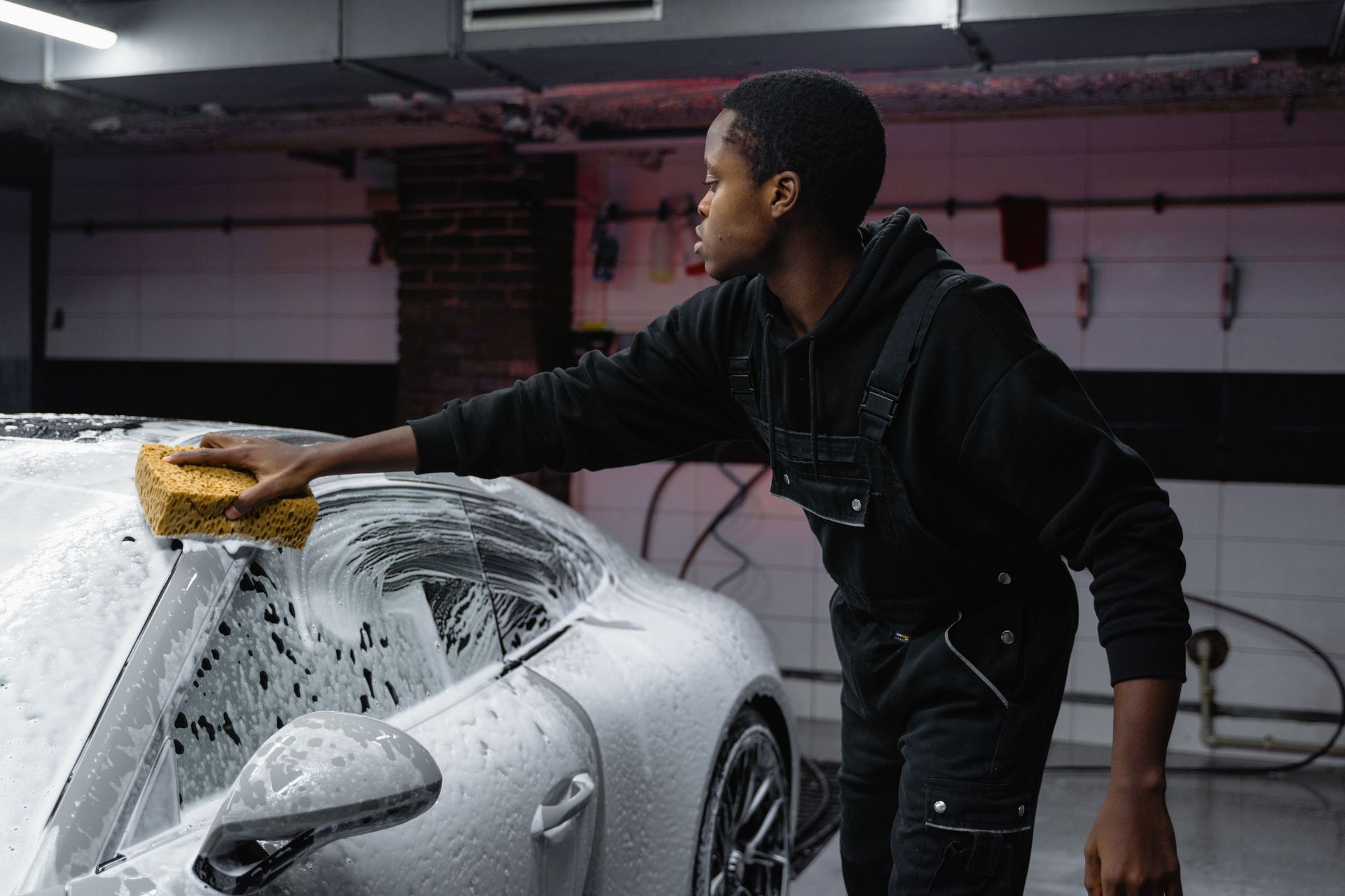 A detailer in a black uniform and cap uses a foam-covered mitt to wash a soap-covered car in a garage.