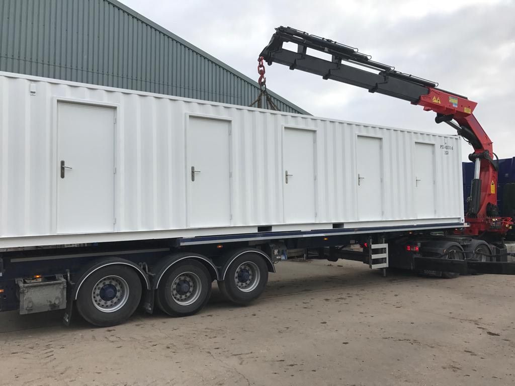 White container with doors loaded on a trailer, being lifted by a crane arm.