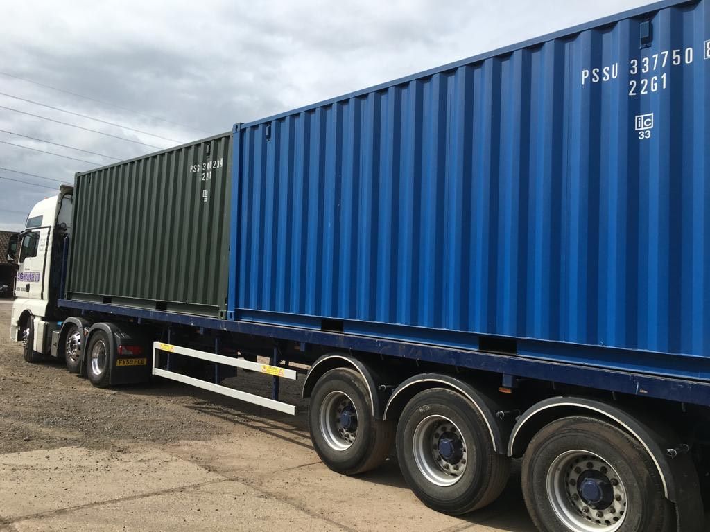 Truck carrying two shipping containers, one green and one blue, against a cloudy sky.