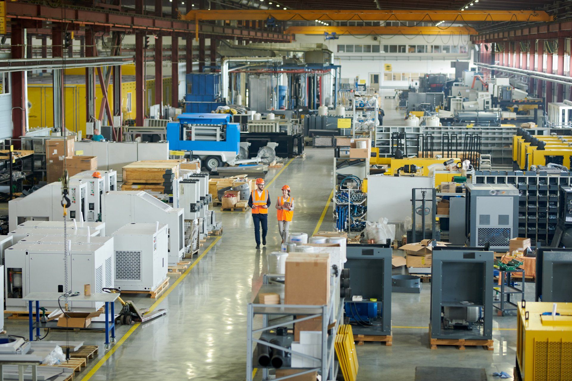 Two workers in safety vests walking through a busy factory floor, surrounded by machinery and equipment.