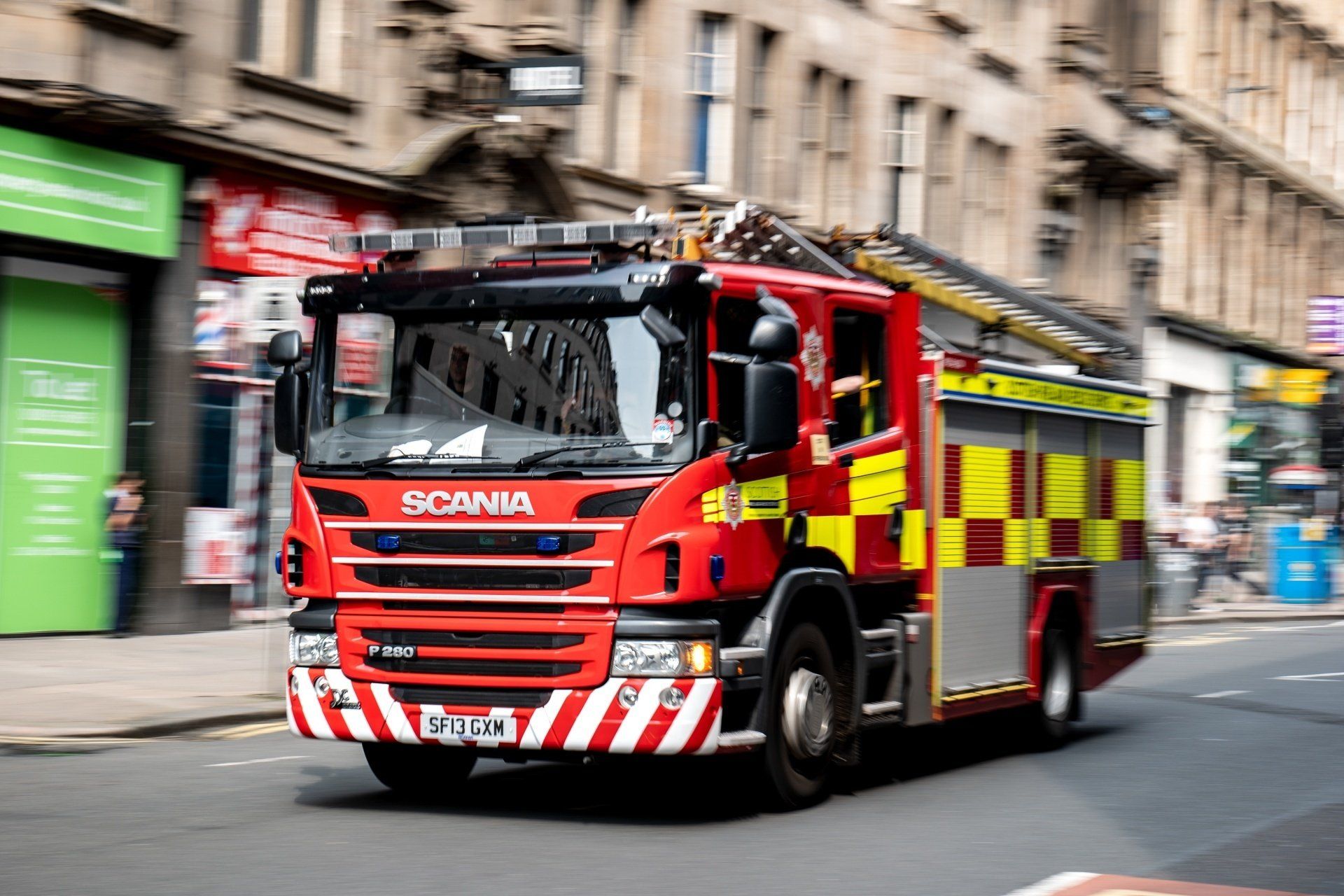 Red fire engine driving on city street, red and yellow checkered panel on side.
