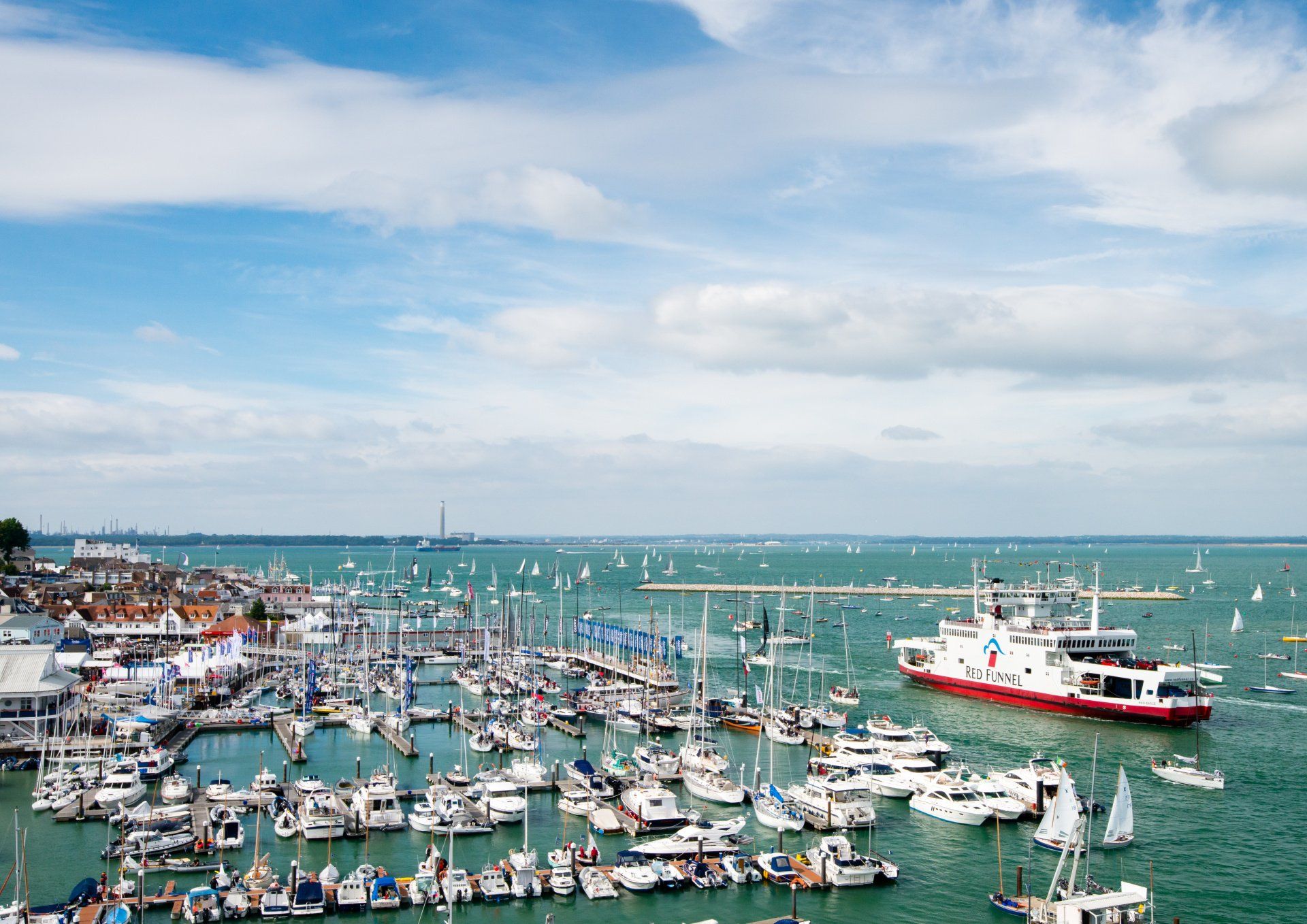 Boats in a harbor under a partly cloudy sky, with a large red and white ship and small buildings along the shore.