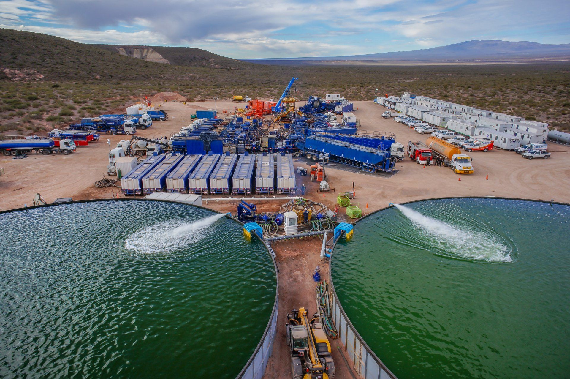 Oil well site with multiple trucks and tanks, two water basins, and equipment in a desert landscape.
