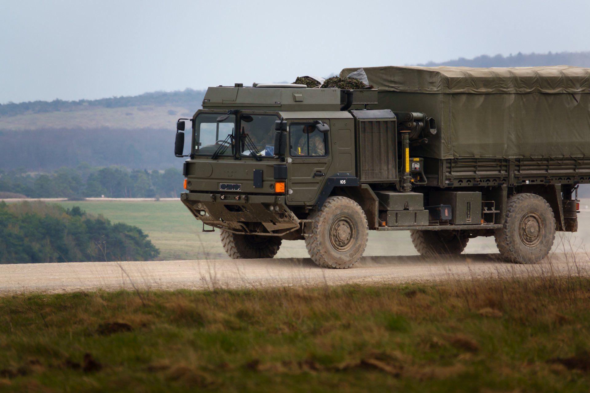 Green military truck driving on a dirt road, with a field and hills in the background.
