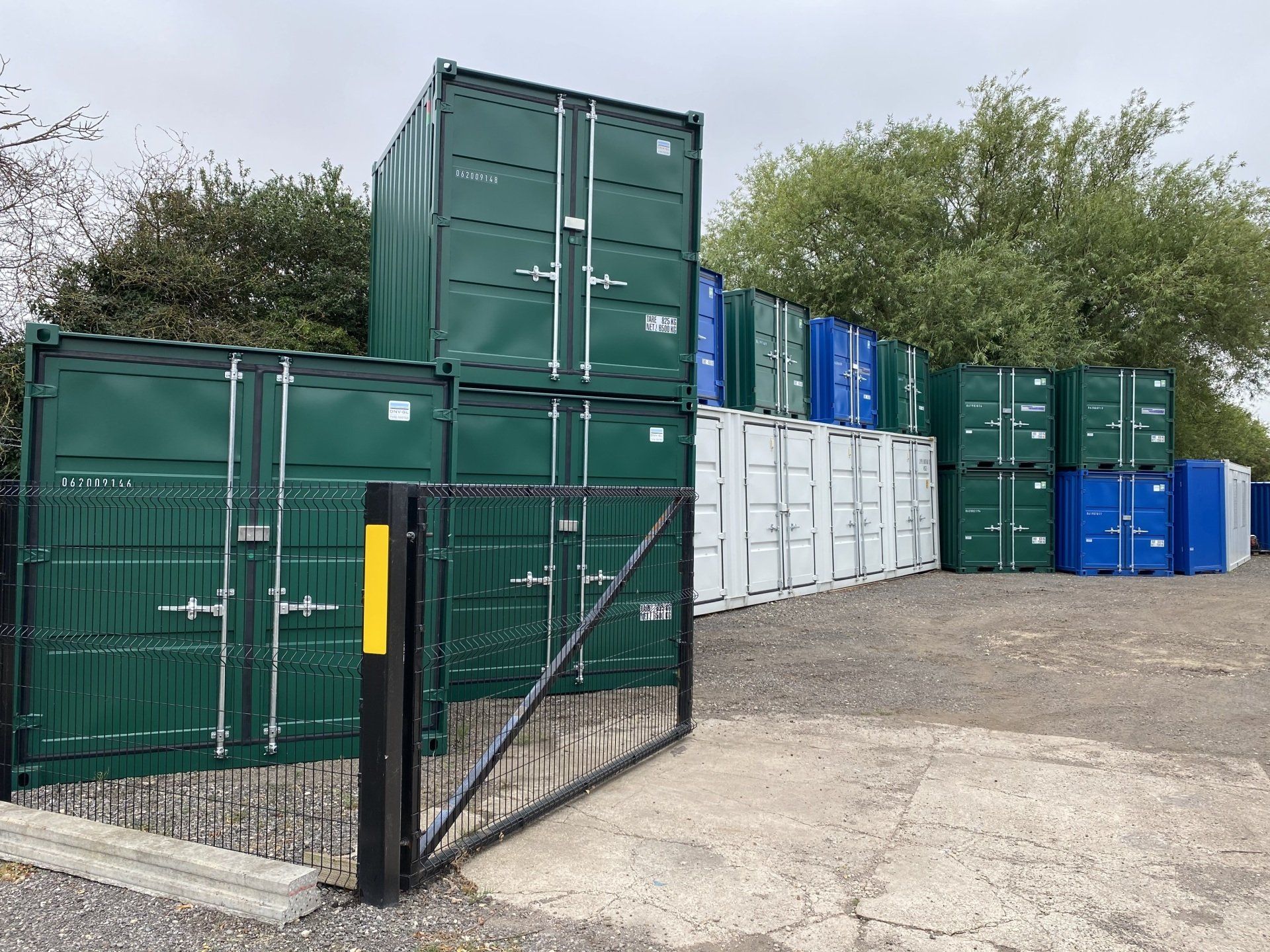 Storage containers behind a black metal gate, in various colors, on a gravel lot.