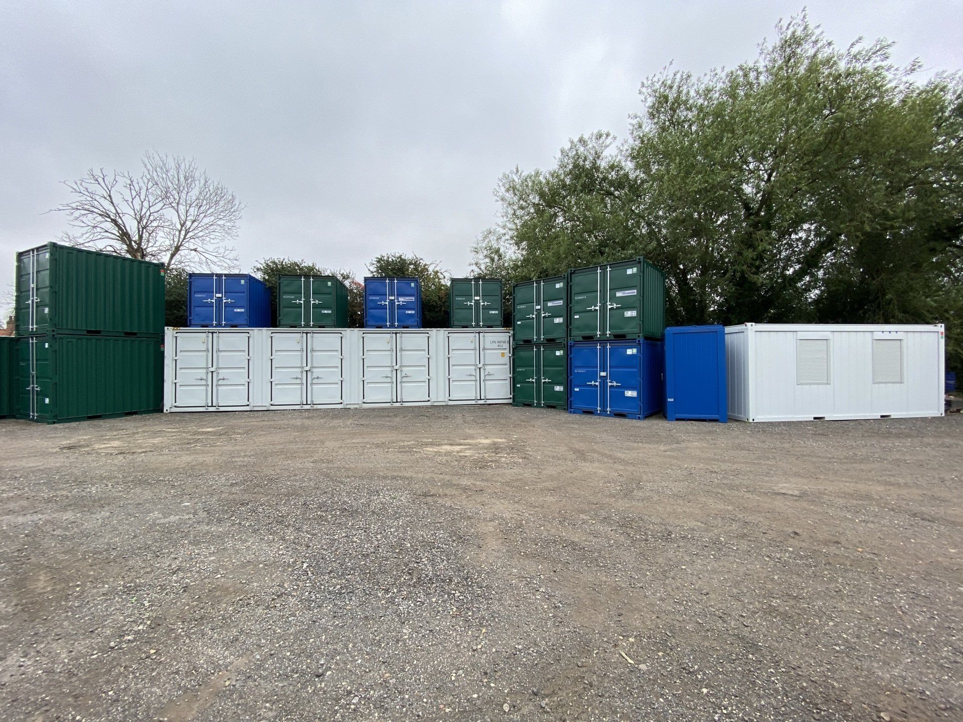 Storage containers of various colors, including green, blue, and white, sit on a gravel surface under a cloudy sky.