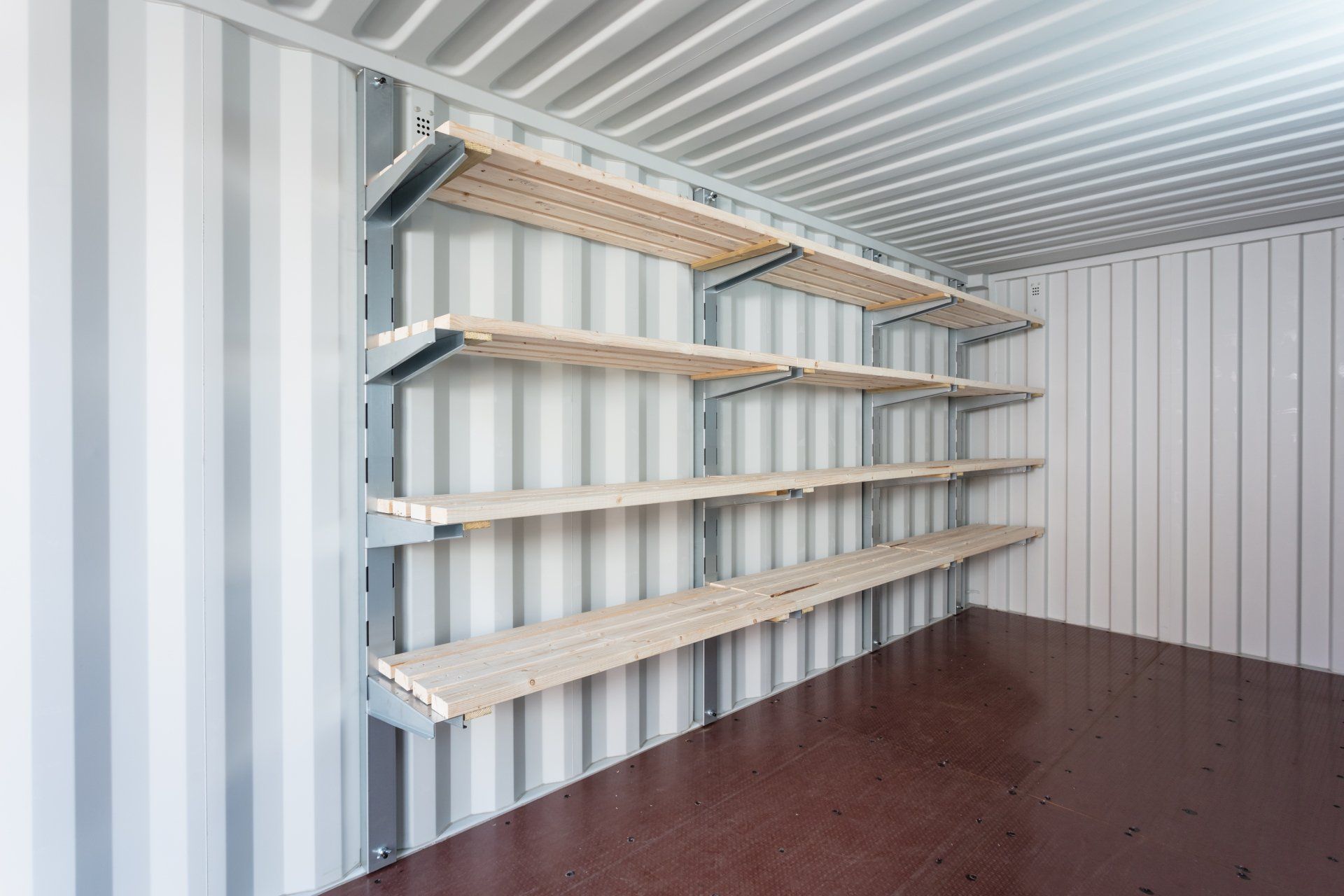 Interior of a metal storage container with shelving. Gray container walls, wooden shelves, and a brown floor.