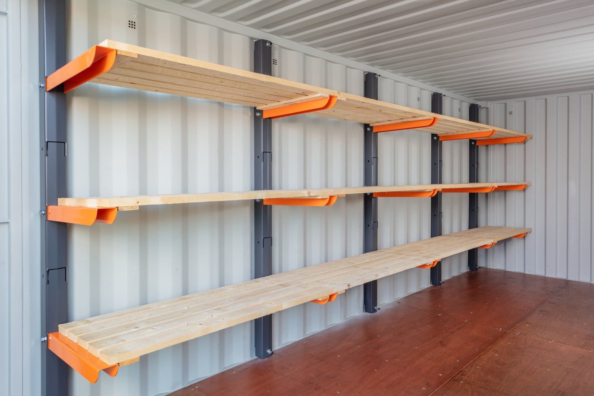 Three wooden shelves with orange brackets attached to a corrugated metal wall inside a storage container.