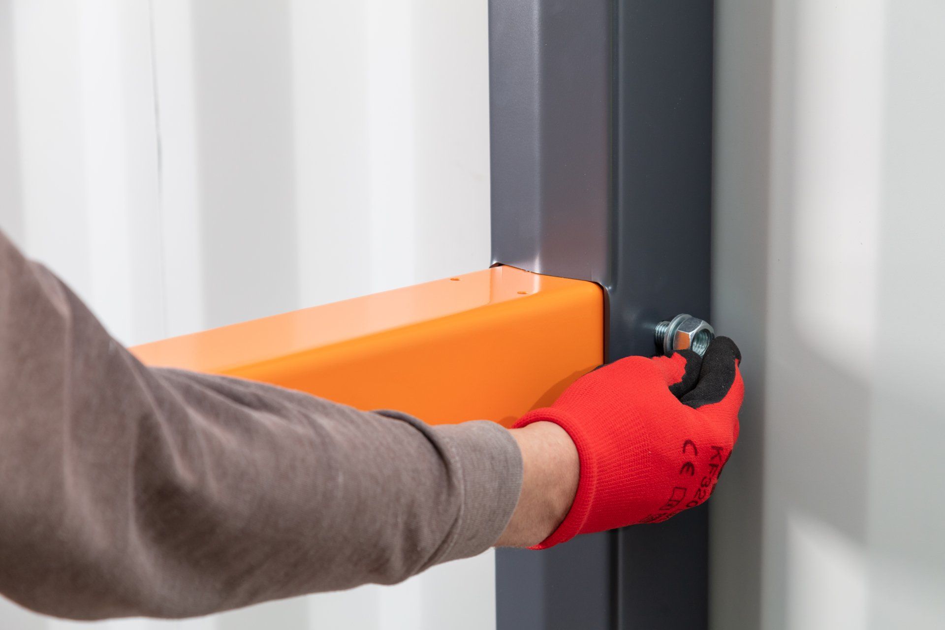 Person wearing a red glove, tightening a bolt on an orange beam attached to a gray metal frame.