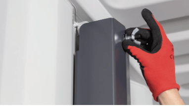 A gloved hand uses a wrench to tighten a bolt on a gray rectangular component, mounted on a white surface.