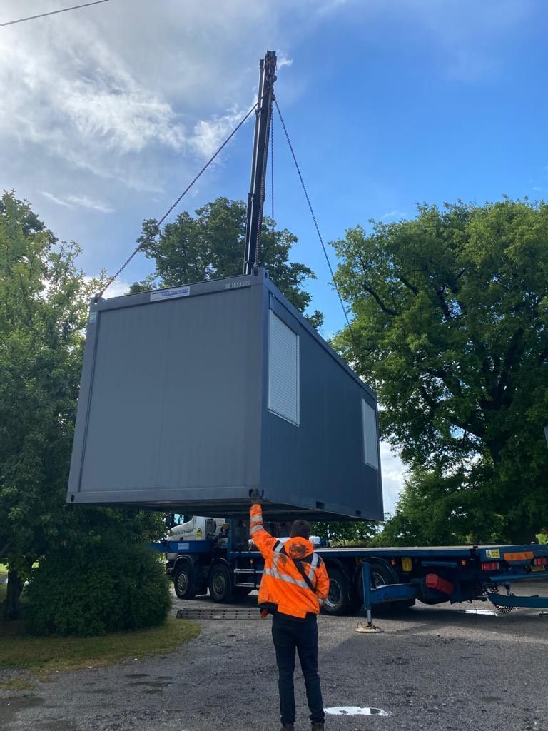 A crane lifting a gray modular building. A person in an orange vest directs the lift. Outdoors, blue sky.