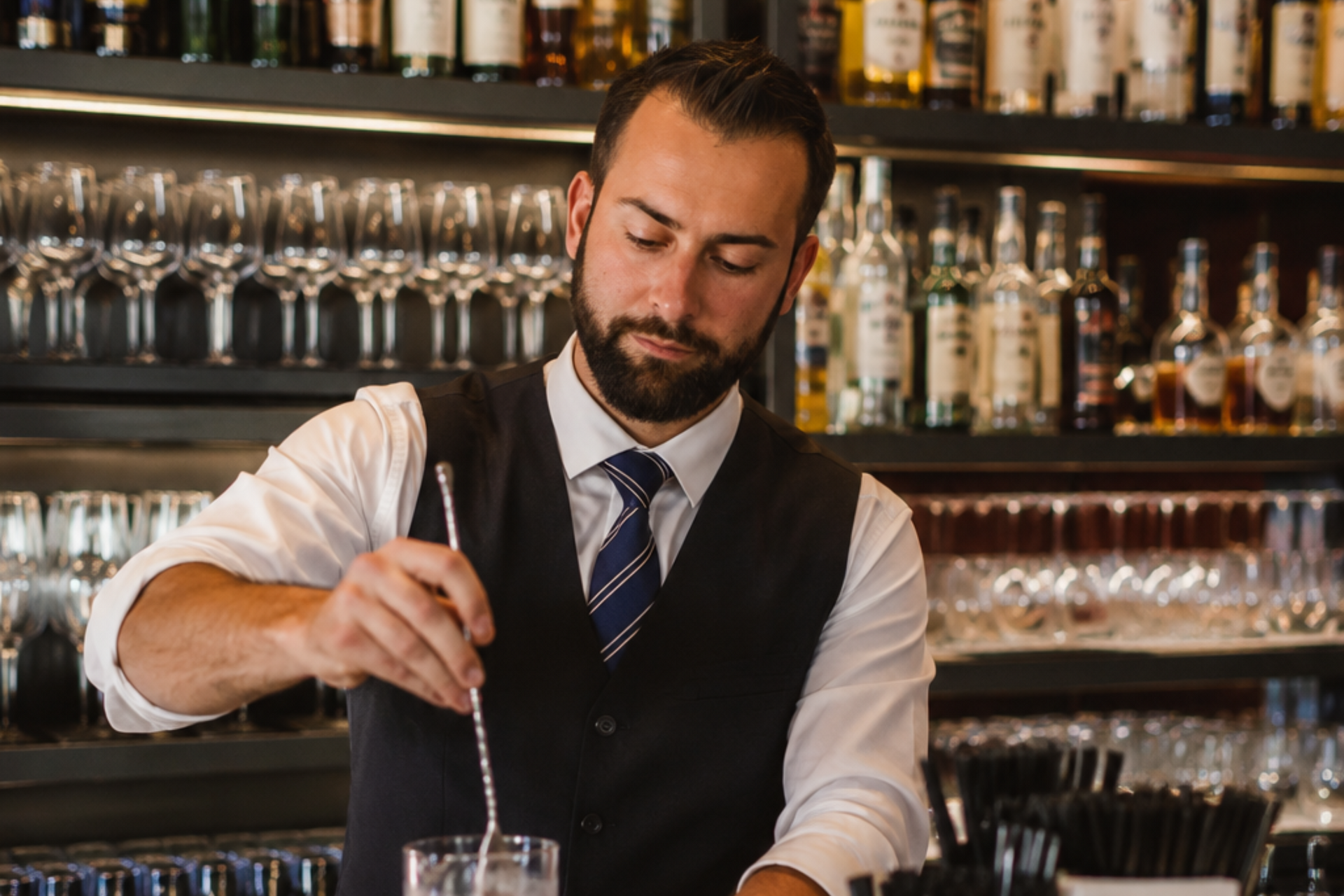 Bartender in vest and tie stirring a drink behind a bar with rows of bottles and glasses.