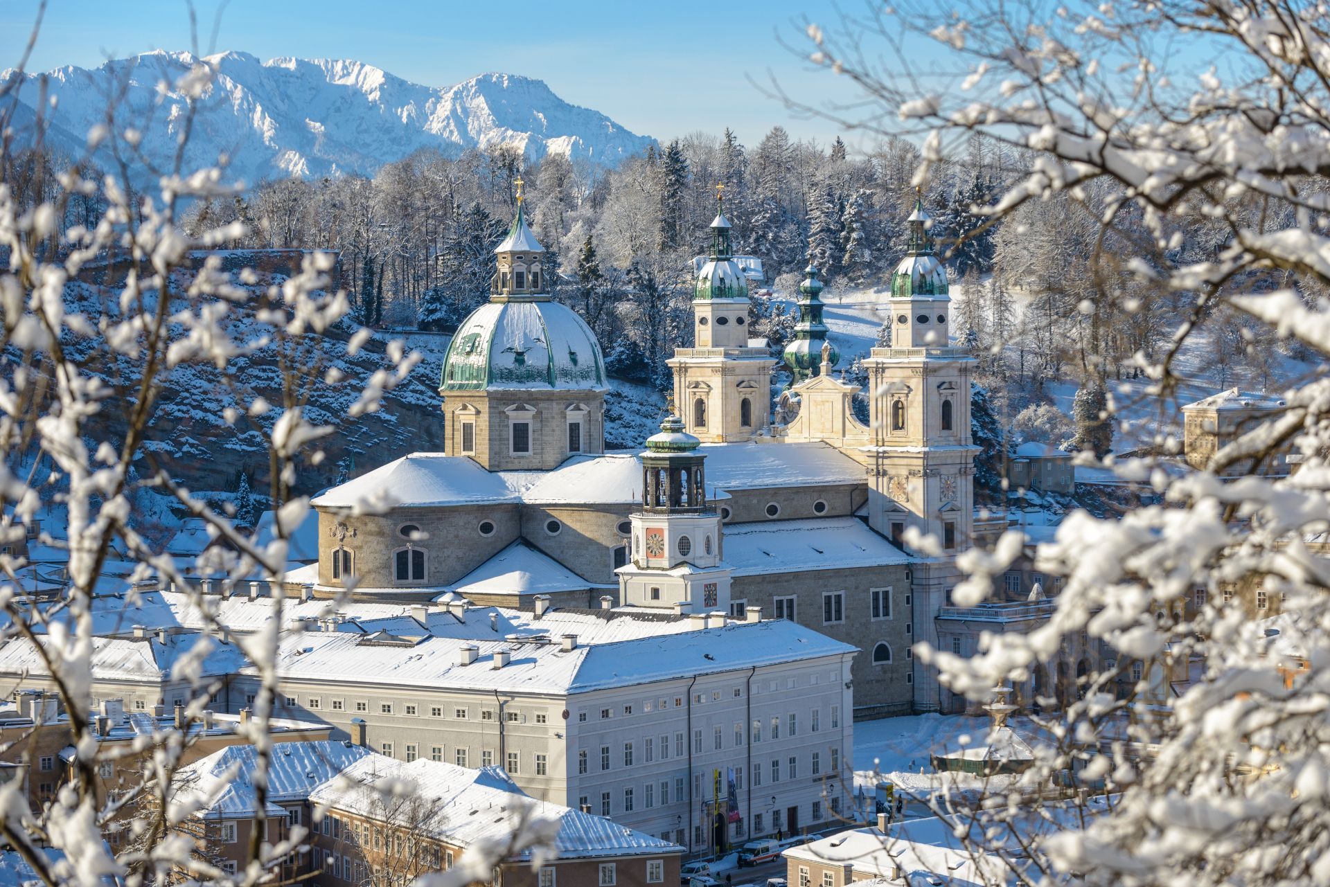Snow-covered Salzburg Cathedral with its iconic green dome, framed by frost-covered tree branches against distant mountains.
