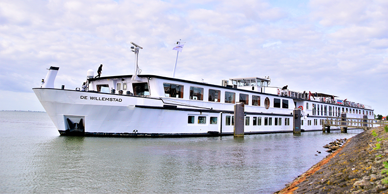White passenger ferry docked on a river beside a rocky shoreline under an overcast sky