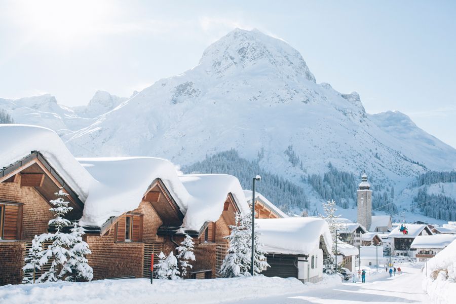 Wooden houses with heavy snow-covered roofs in a sunny, mountainous alpine village with a church tower in the distance.