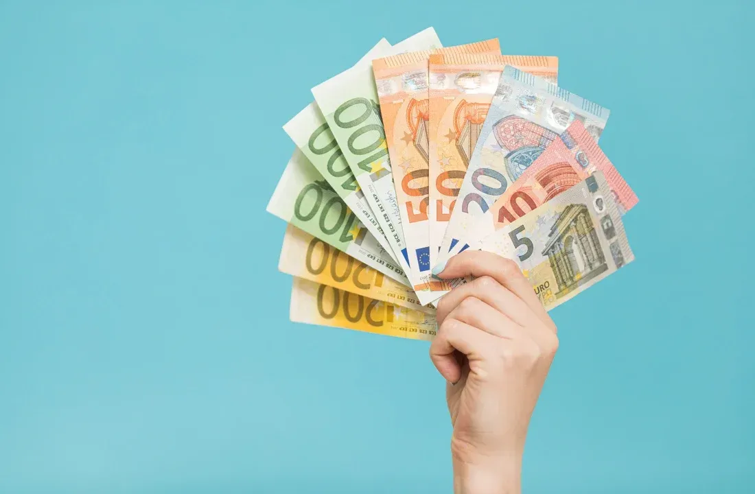 A hand holds a fan of various Euro banknotes against a plain light blue background.
