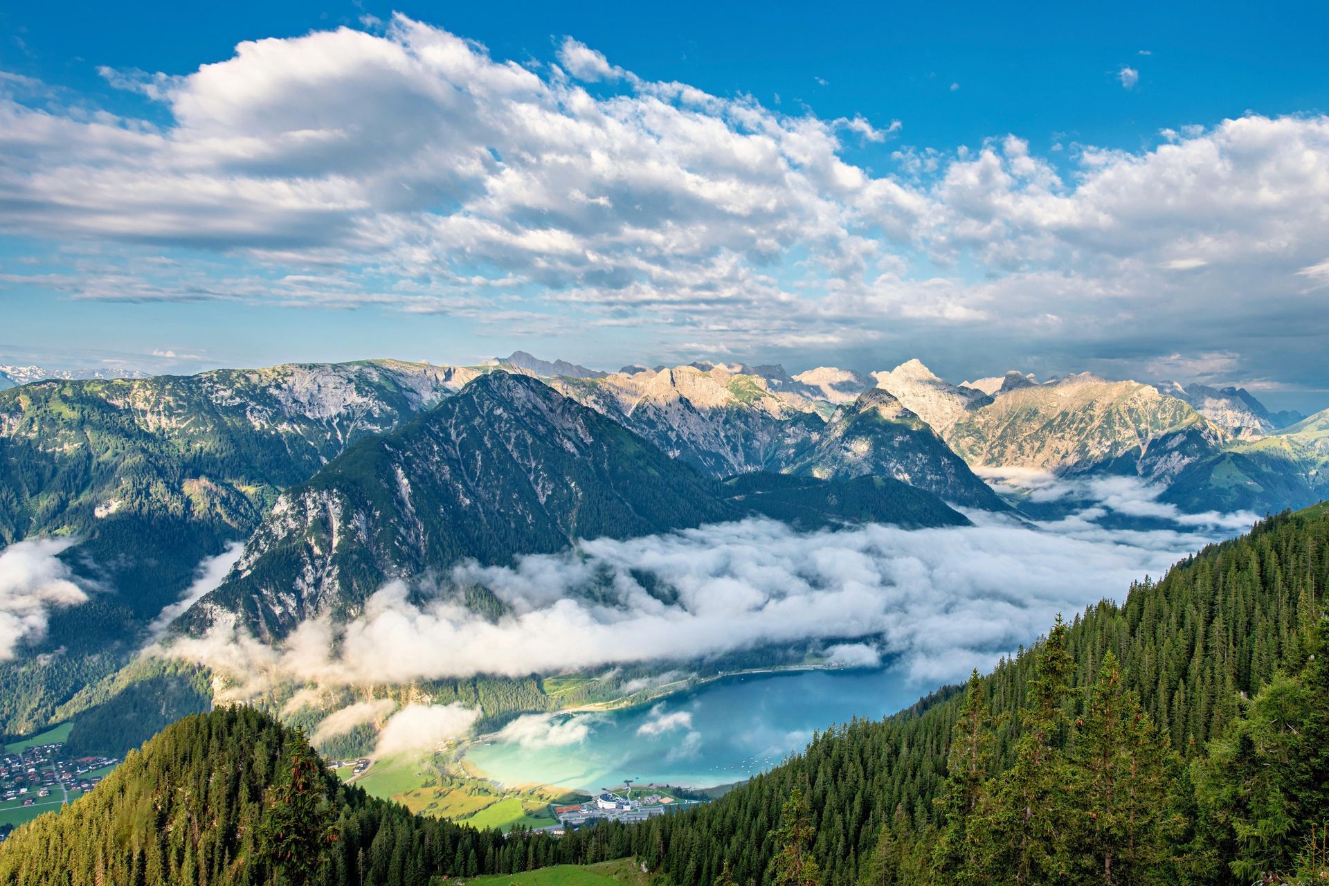A scenic view of a bright blue mountain lake partially obscured by low-hanging white clouds, set against rugged peaks.