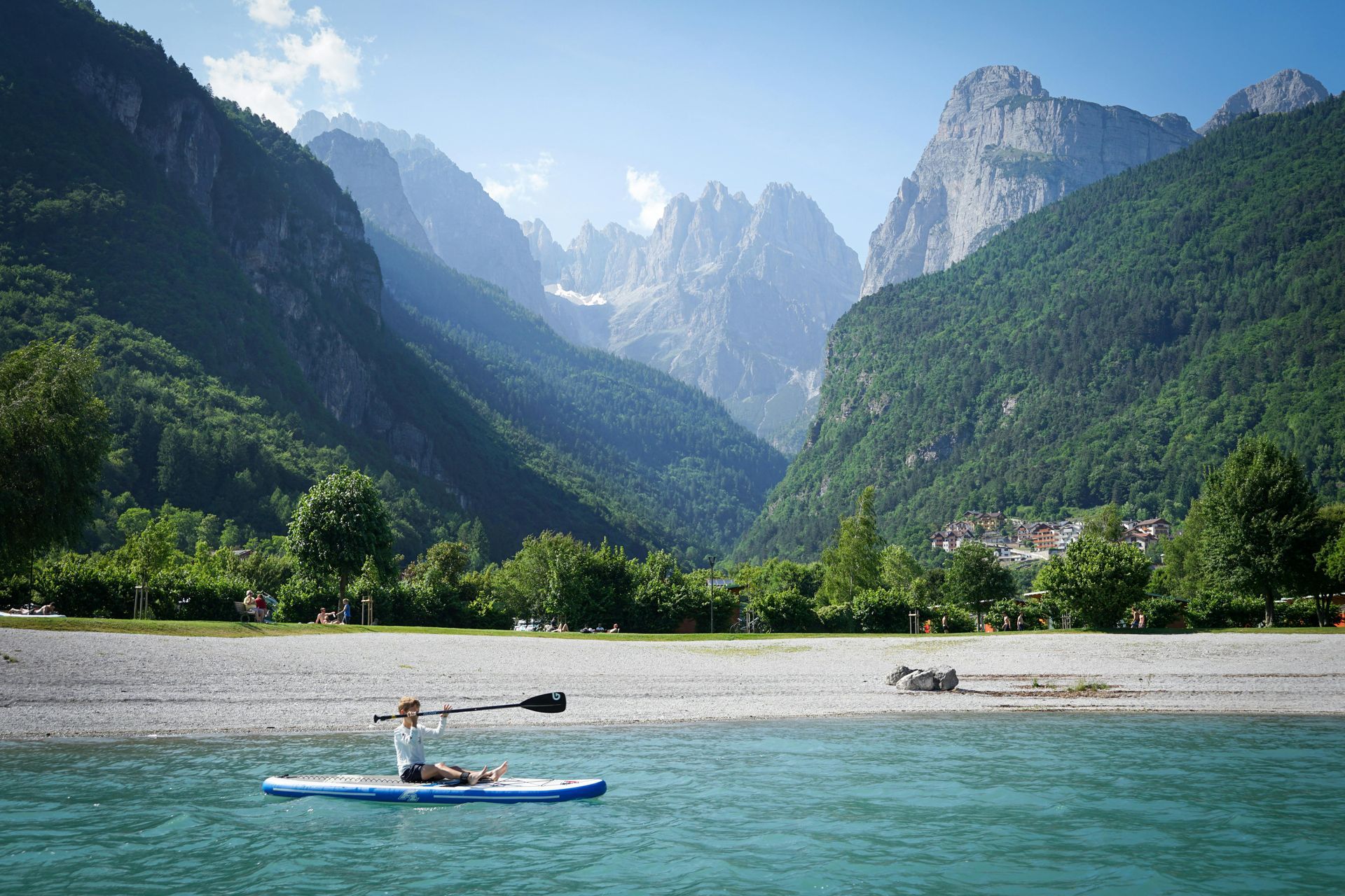 A person paddles on a blue stand-up paddleboard in a turquoise lake before a backdrop of lush green mountains and peaks.