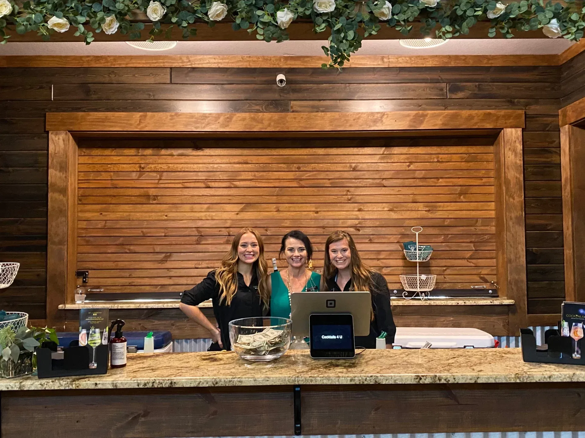 Three women are standing behind a counter in front of a wooden wall.
