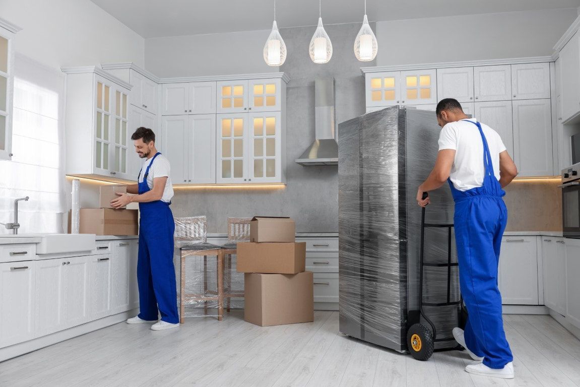 Two movers packing boxes and moving a wrapped refrigerator in a white kitchen.