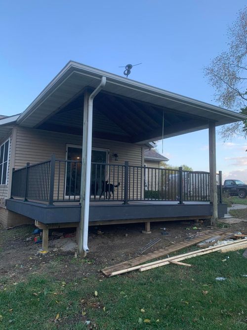 A grey deck with black railings under a covered porch roof, attached to a house on a grassy lot.