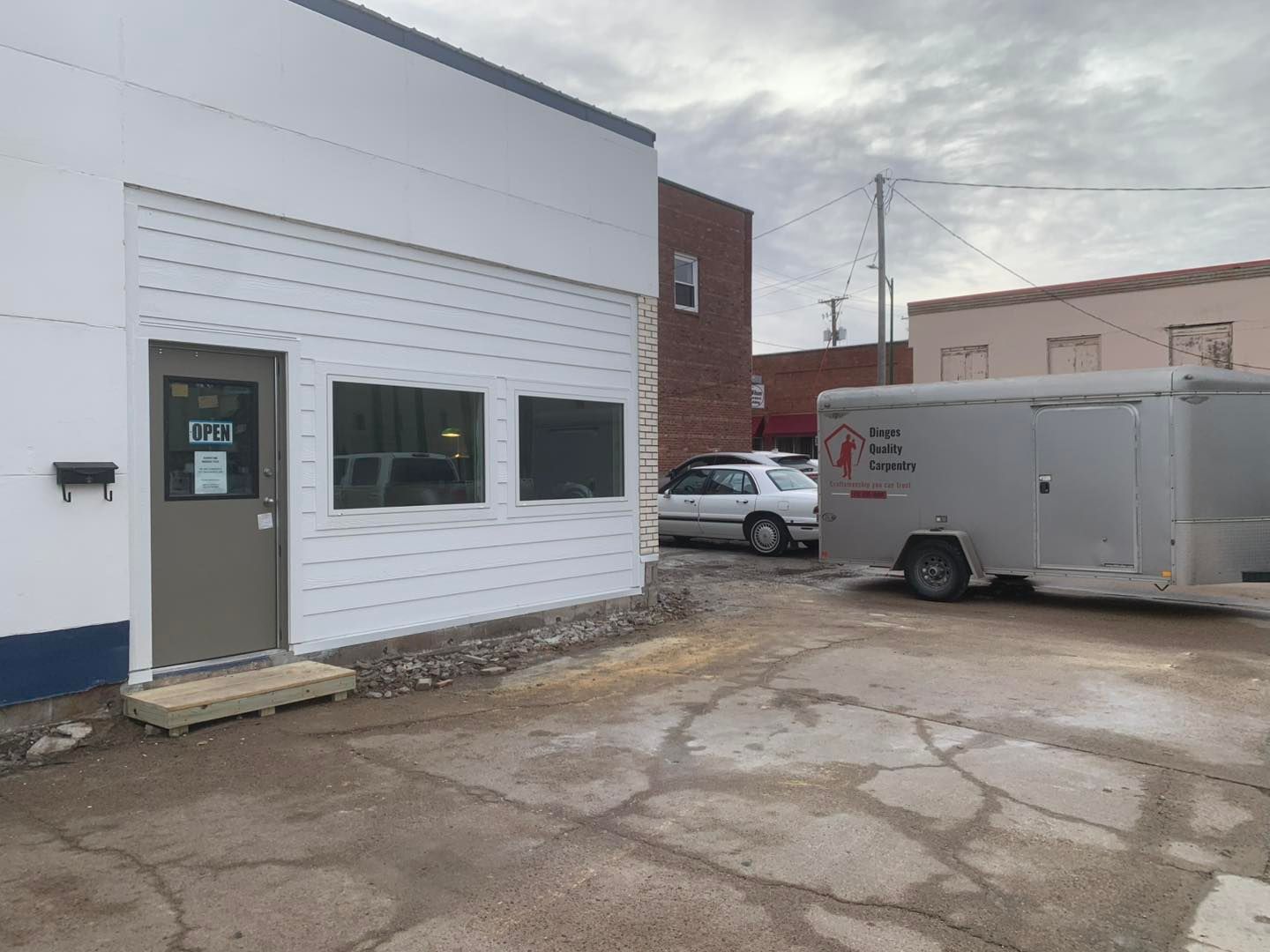 A white building exterior with a tan door, two windows, and a silver cargo trailer parked in a gravel lot.