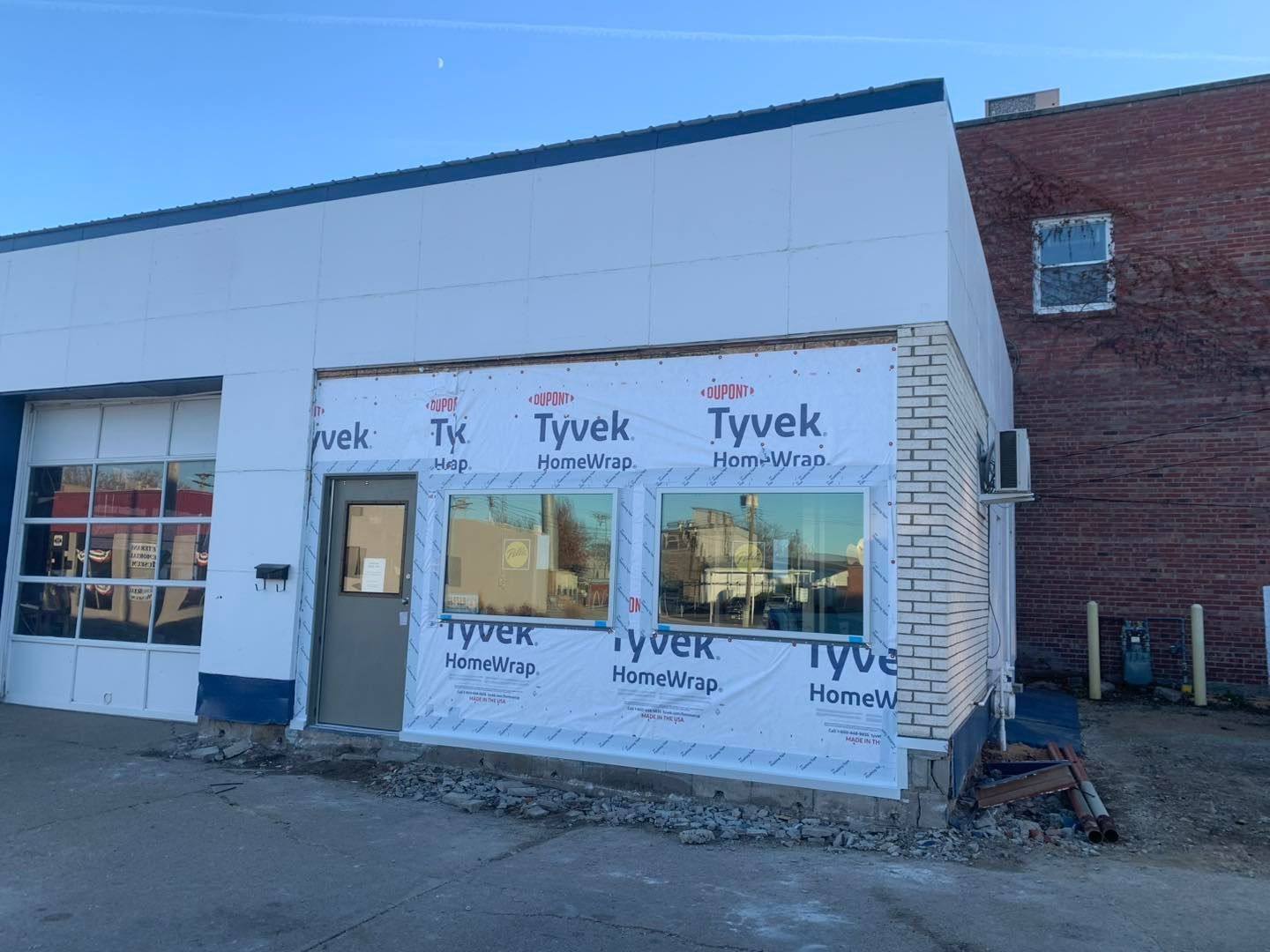 A white commercial building undergoing renovations, with Tyvek house wrap visible on its front wall next to a glass door.