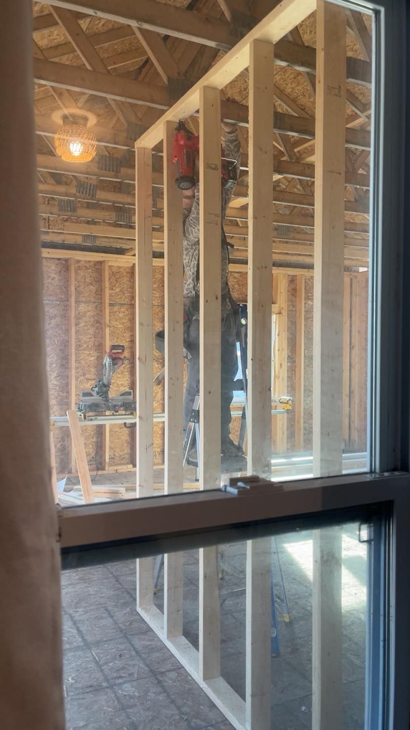 A construction worker framing an interior wall inside a partially finished wooden building, seen through a window.
