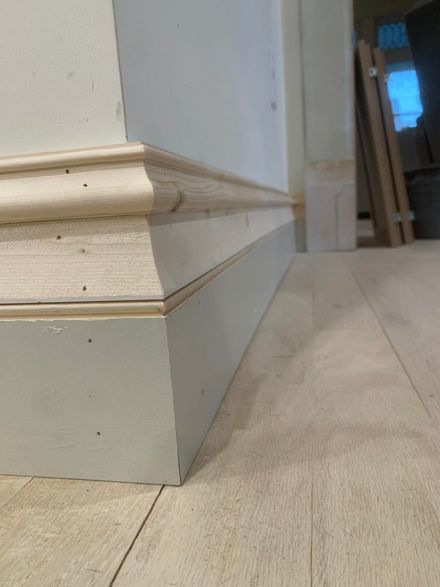 A close-up view of a multi-layered, unfinished wooden baseboard installed against a light-colored wall and wood floor.