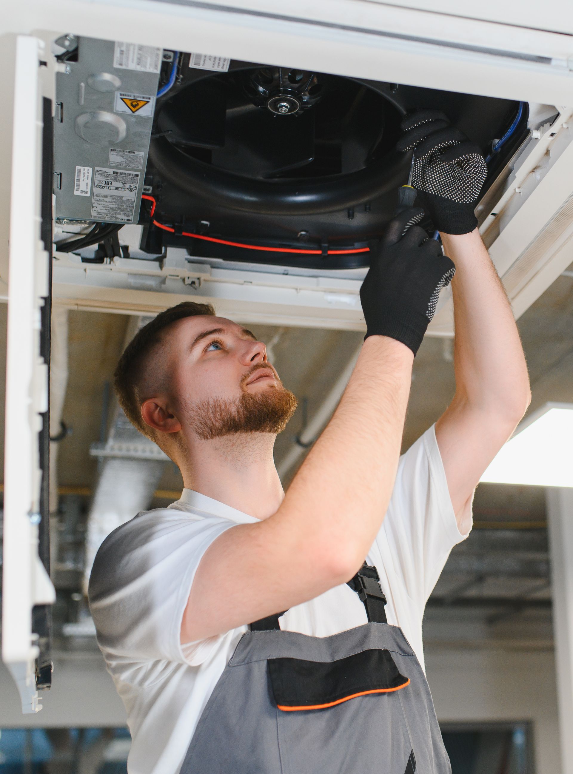 Technician replacing a ceiling HVAC unit filter while standing on a ladder, wearing gloves and overalls