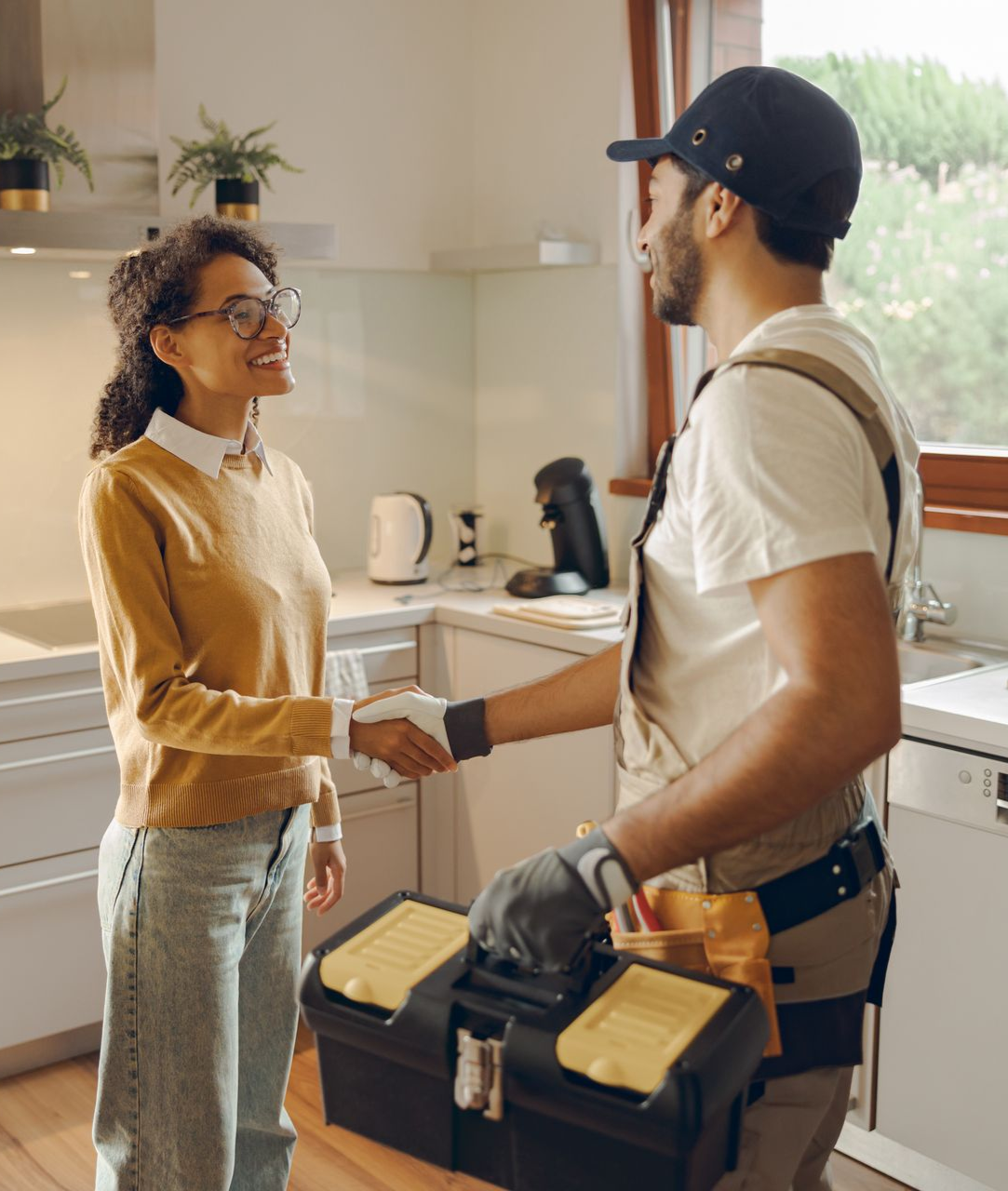 Two people shake hands in a bright kitchen, one carrying a tool bag, suggesting a home repair visit