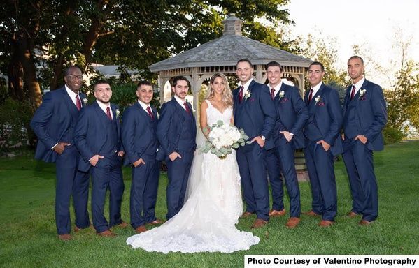 Wedding party posing outdoors in navy suits and a white gown in front of a stone building